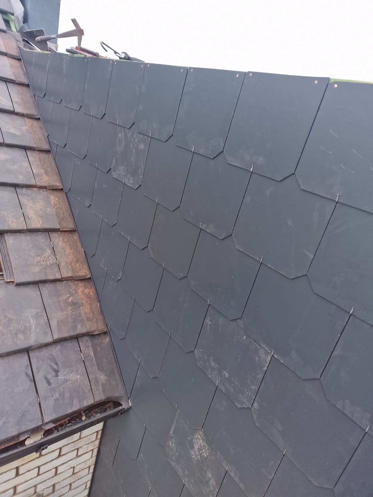 Fragment dachu z nowym, ciemnoszarym pokryciem łupkowym, kontrastujący z fragmentem starego, brązowego pokrycia. Widoczne narzędzia dekarskie na górze dachu.