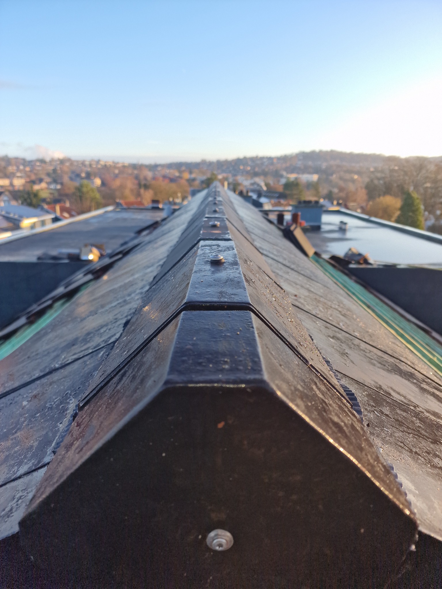 Szczegółowe ujęcie kalenicy dachu pokrytego ciemną dachówką, z widocznym mocowaniem. W tle rozmyty krajobraz miejski pod błękitnym niebem.