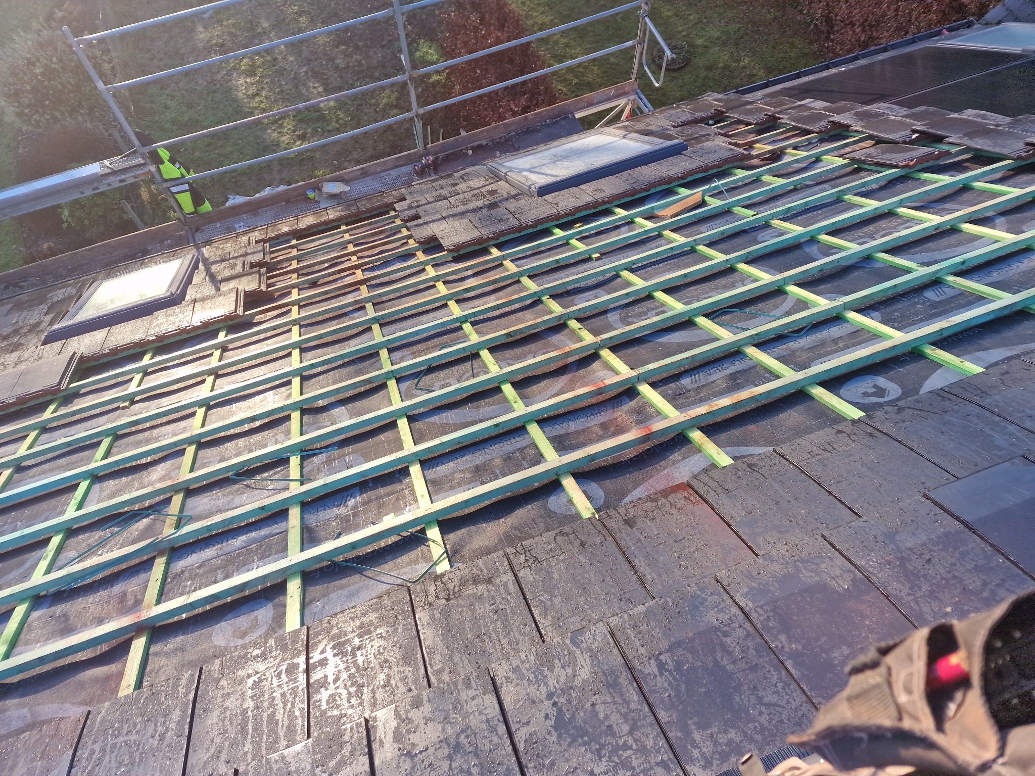 Częściowo pokryty dach z widocznymi łatami i oknami dachowymi, fragment rusztowania w tle. Prace dekarskie w toku, widok z góry na konstrukcję dachu.