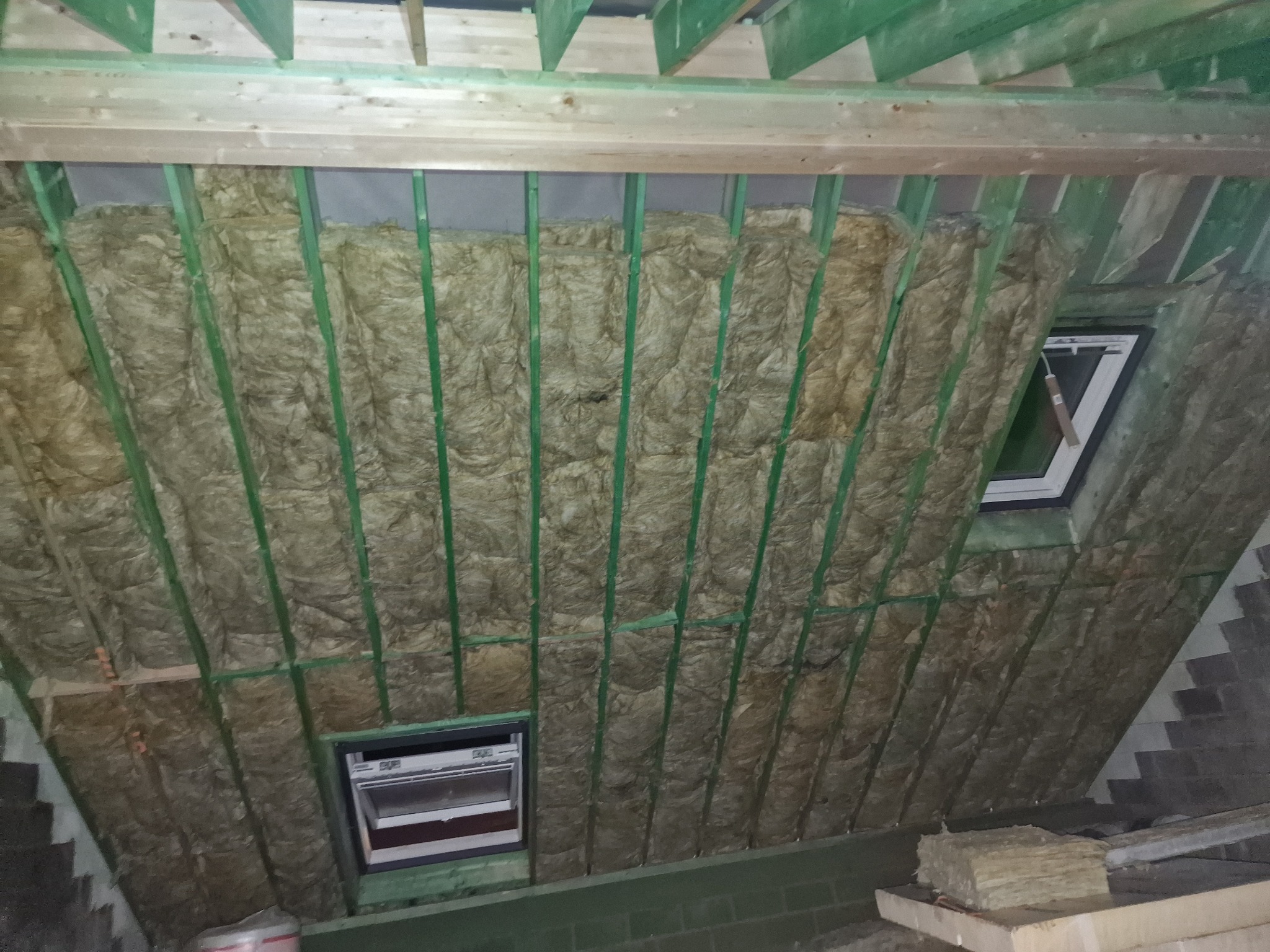 Ocieplone wełną mineralną poddasze z oknami dachowymi, widoczne zielone elementy konstrukcji drewnianej. Częściowo widoczne kostki wełny mineralnej.