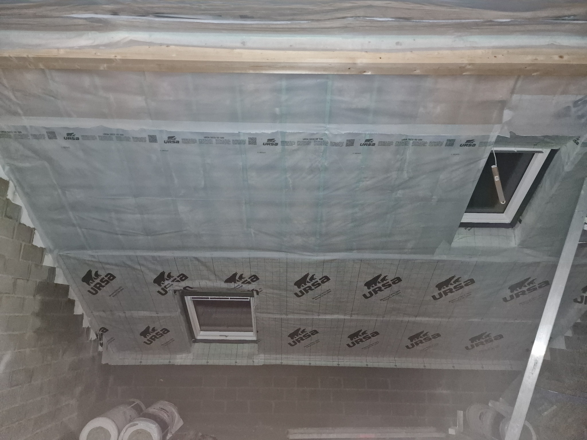 Poddasze w trakcie ocieplania, widoczna folia paroizolacyjna URSA z nadrukowanym logo, okna dachowe i fragment ściany z bloczków. Prace izolacyjne w toku.