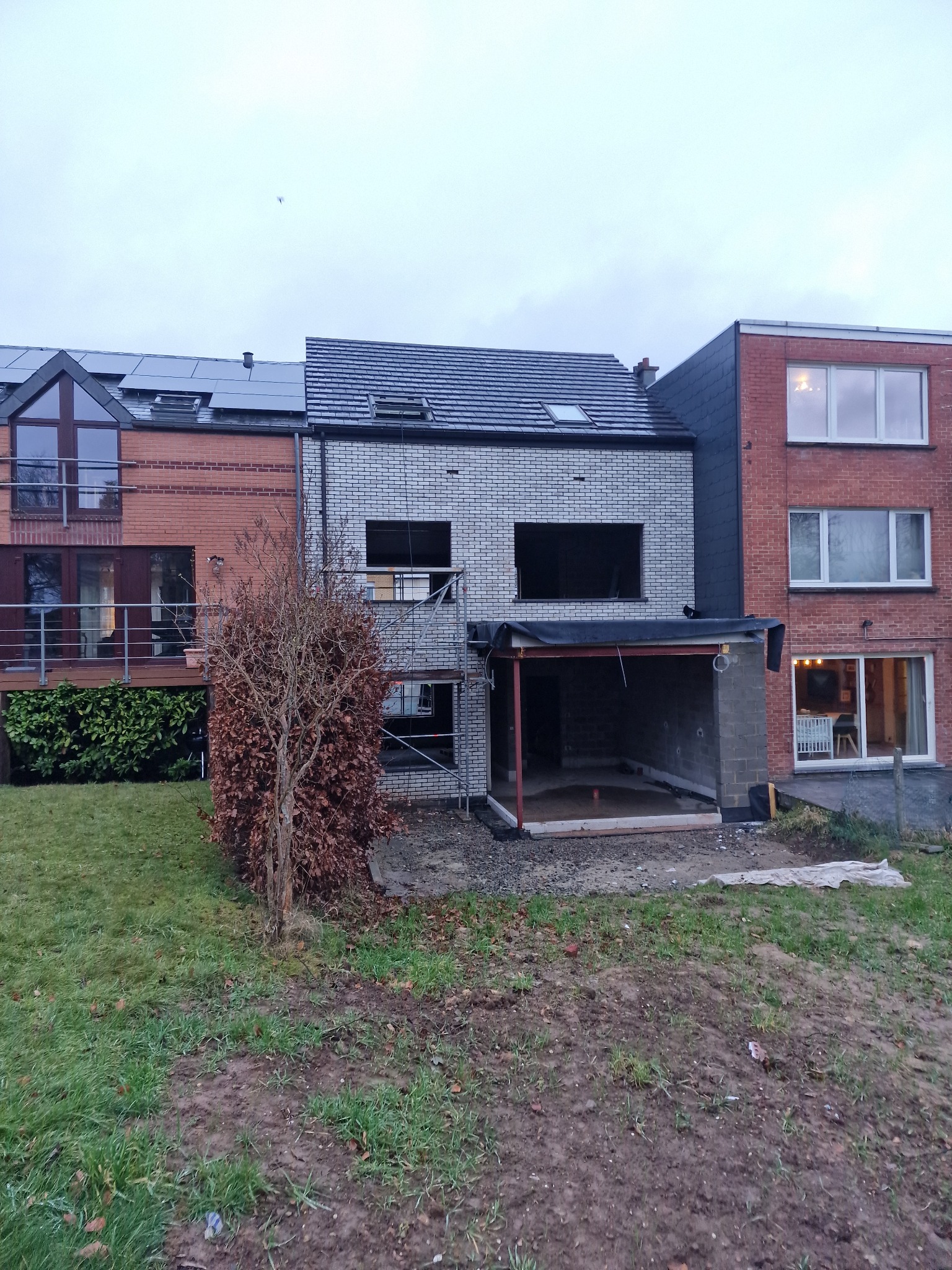 Budynek w trakcie rozbudowy z szarej cegły, widoczne rusztowania i zadaszenie nad dobudowywaną częścią. Obok budynki z czerwonej cegły i zielony trawnik.