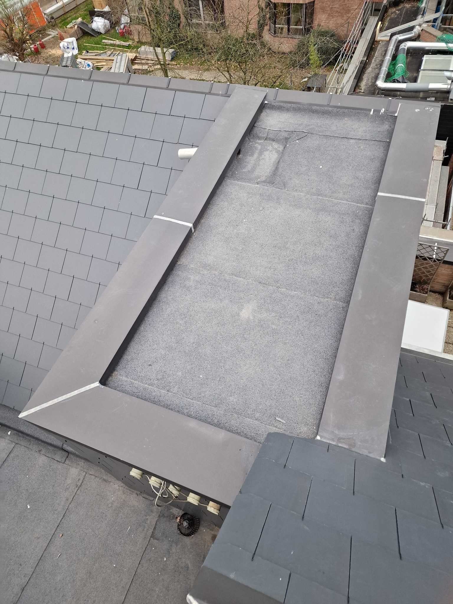 Szary dach z wykończonym obramowaniem lukarny i fragmentem papy. Widok z góry ukazuje detale pokrycia i obróbki blacharskiej.