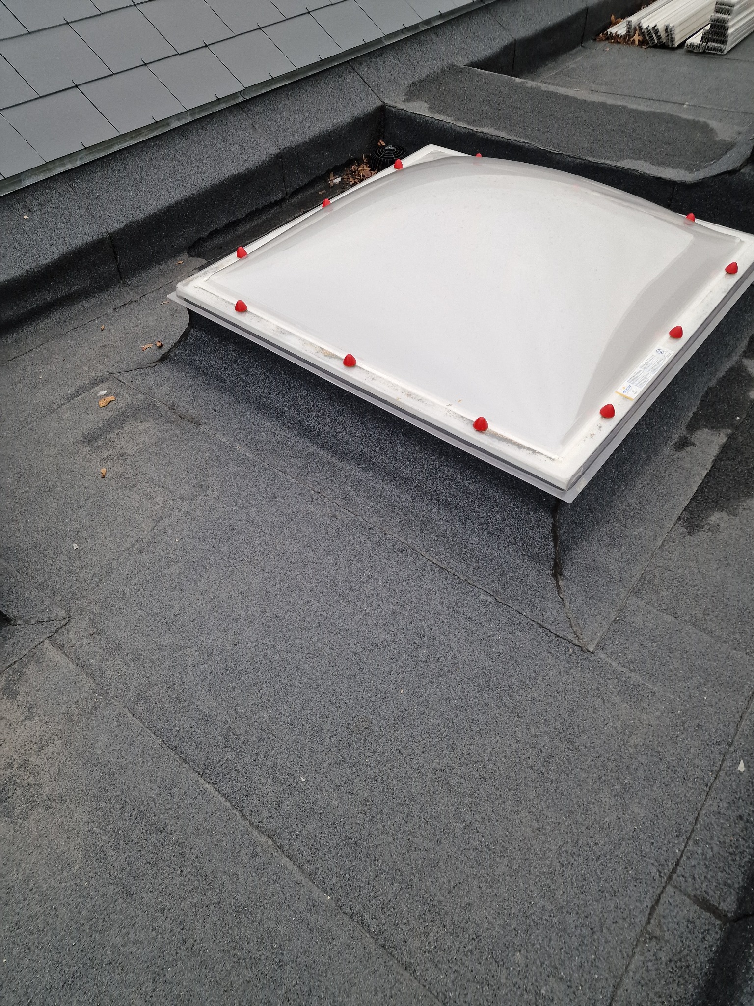 Płaski dach pokryty papą z zamontowanym świetlikiem dachowym z czerwonymi zaślepkami. W tle widoczne dachówki i elementy montażowe.