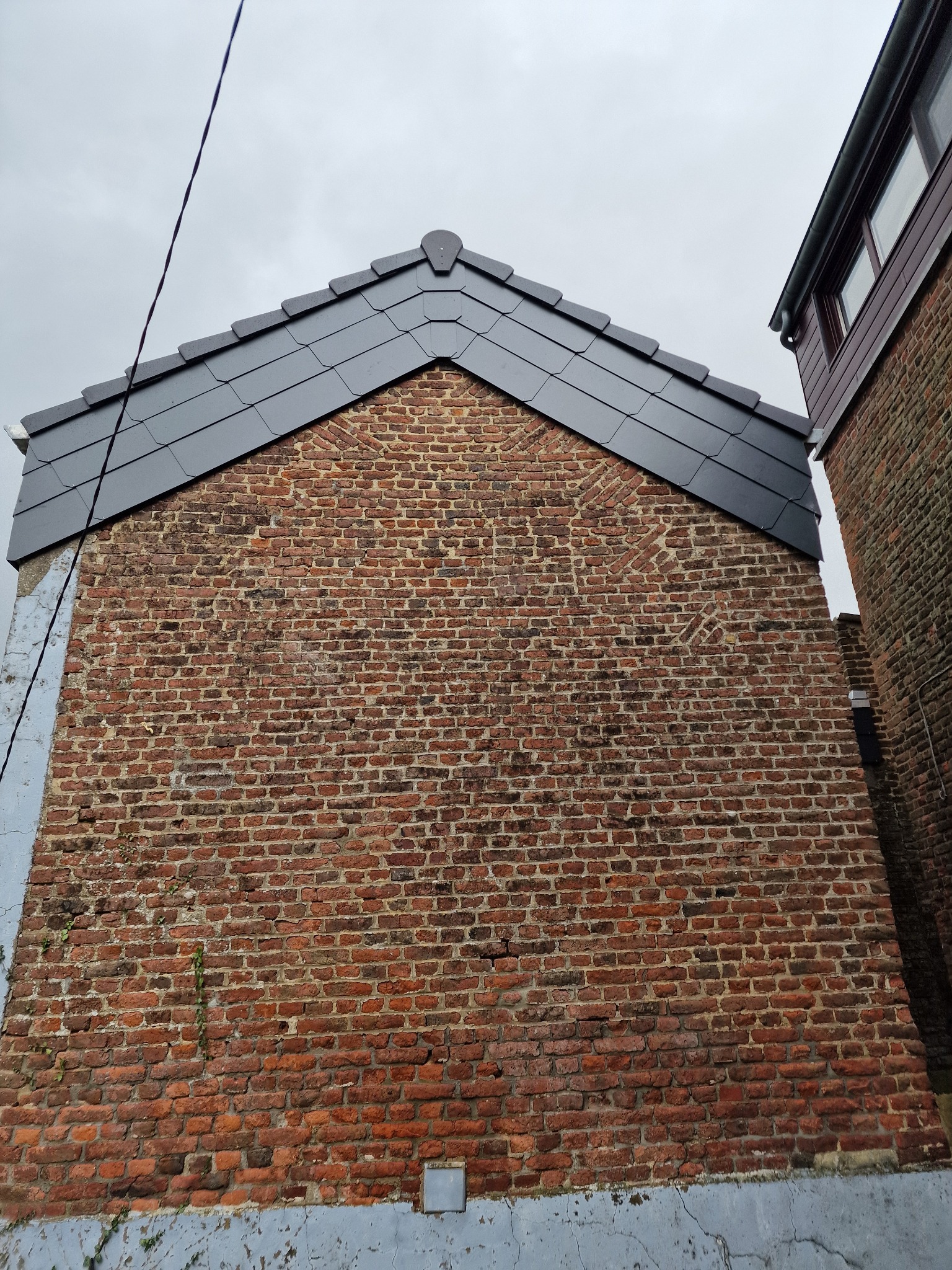 Murowana ściana szczytowa z nowym, grafitowym pokryciem dachowym z dachówki ceramicznej. Widoczne detale wykończenia i sąsiedni budynek z rynną.