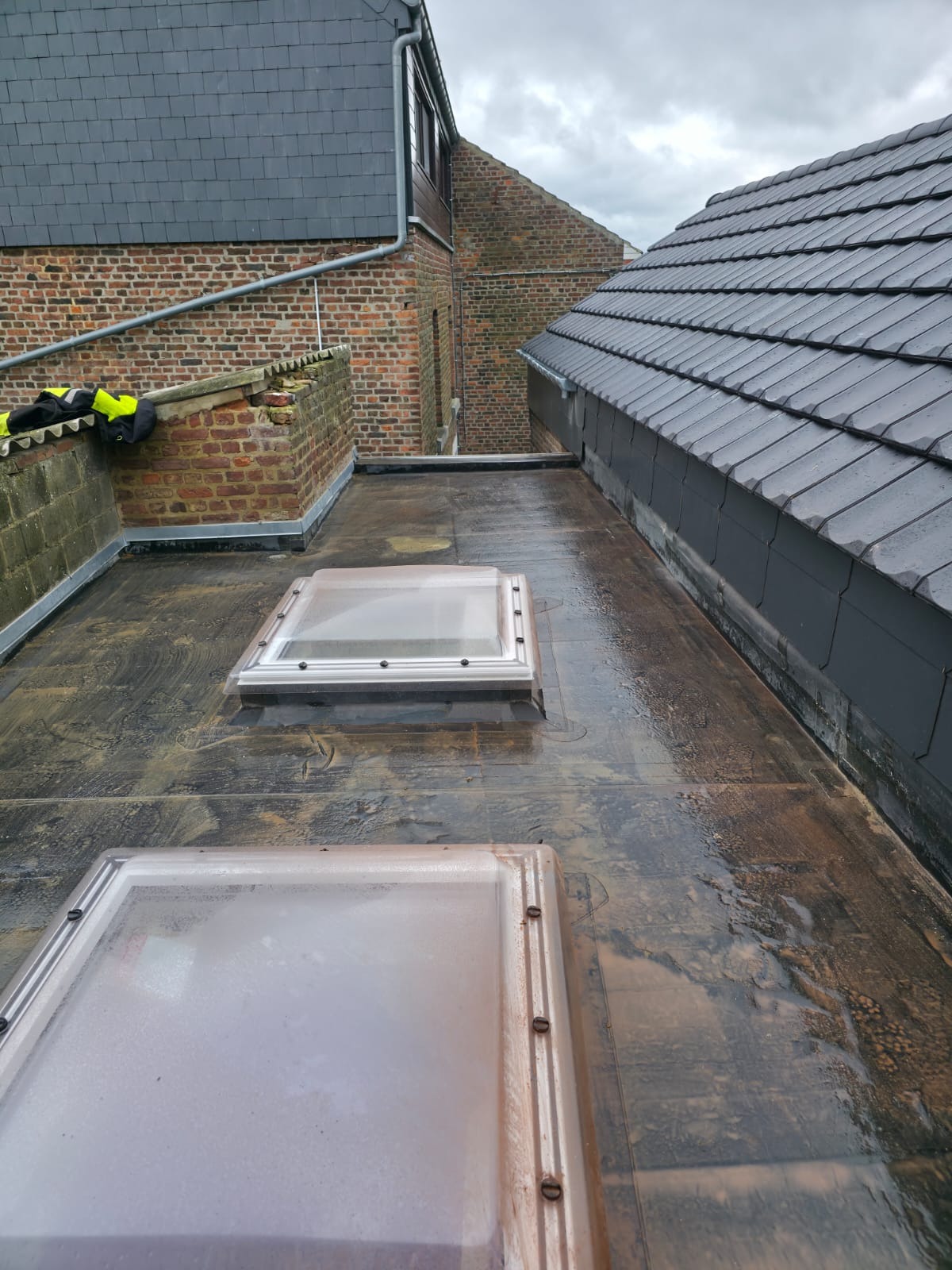 Płaski dach pokryty papą z dwoma świetlikami. Widoczne są sąsiednie budynki z cegły i dachówki. Dach jest mokry, prawdopodobnie po deszczu.