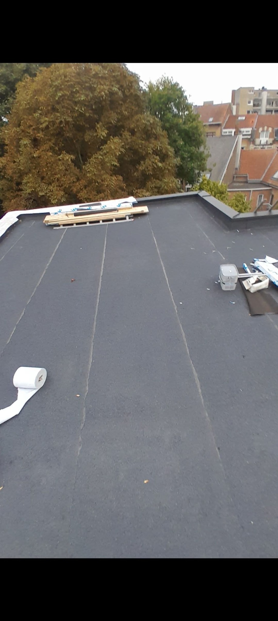 Czarny, płaski dach pokryty papą, z widocznymi narzędziami i materiałami budowlanymi. W tle drzewa i budynki mieszkalne. Widok z góry, w trakcie prac dekarskich.