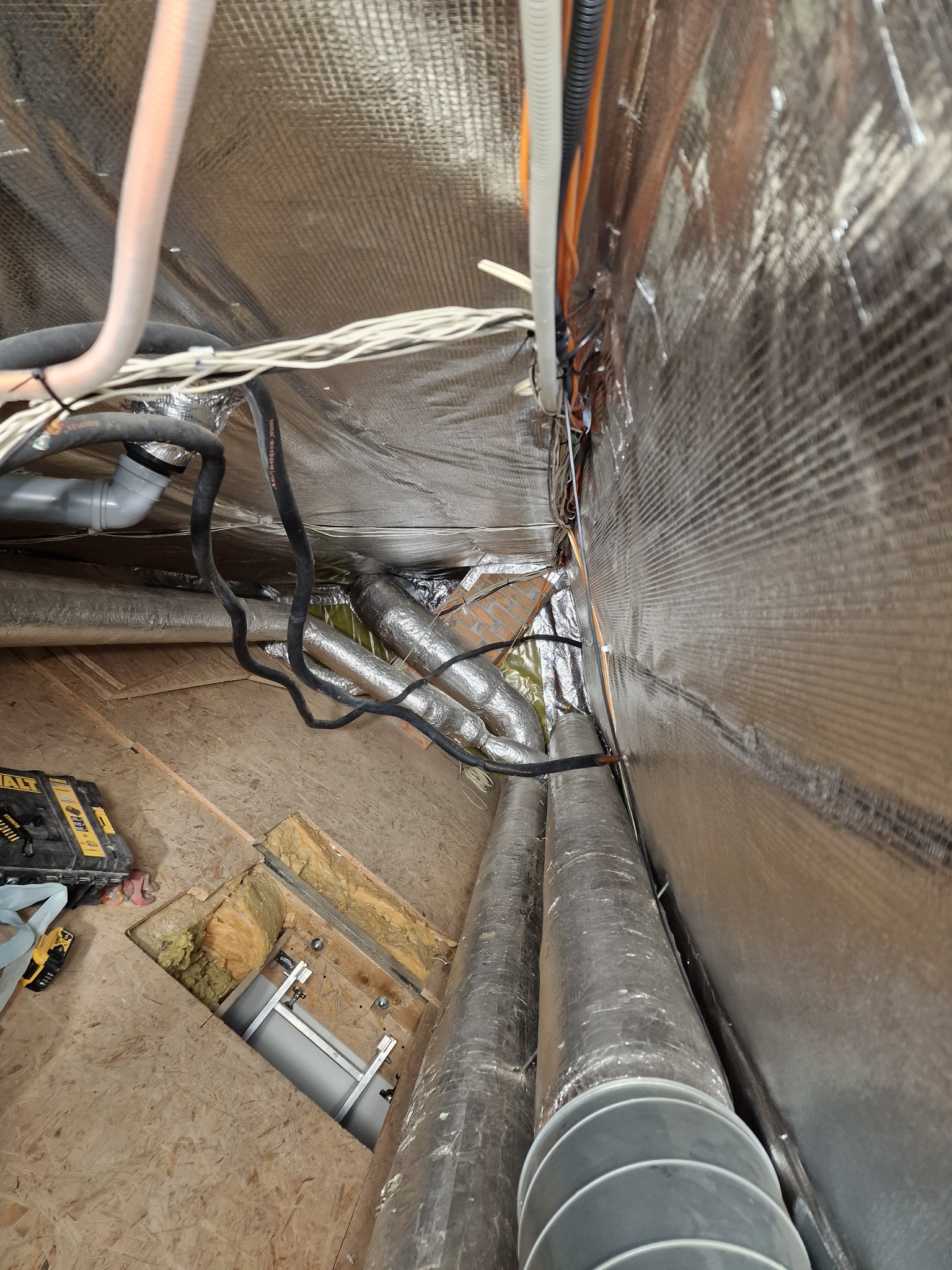 Poddasze z izolacją folią aluminiową, widoczne rury wentylacyjne, kable i narzędzia. Fragment ocieplenia wełną mineralną. Ujęcie z góry.