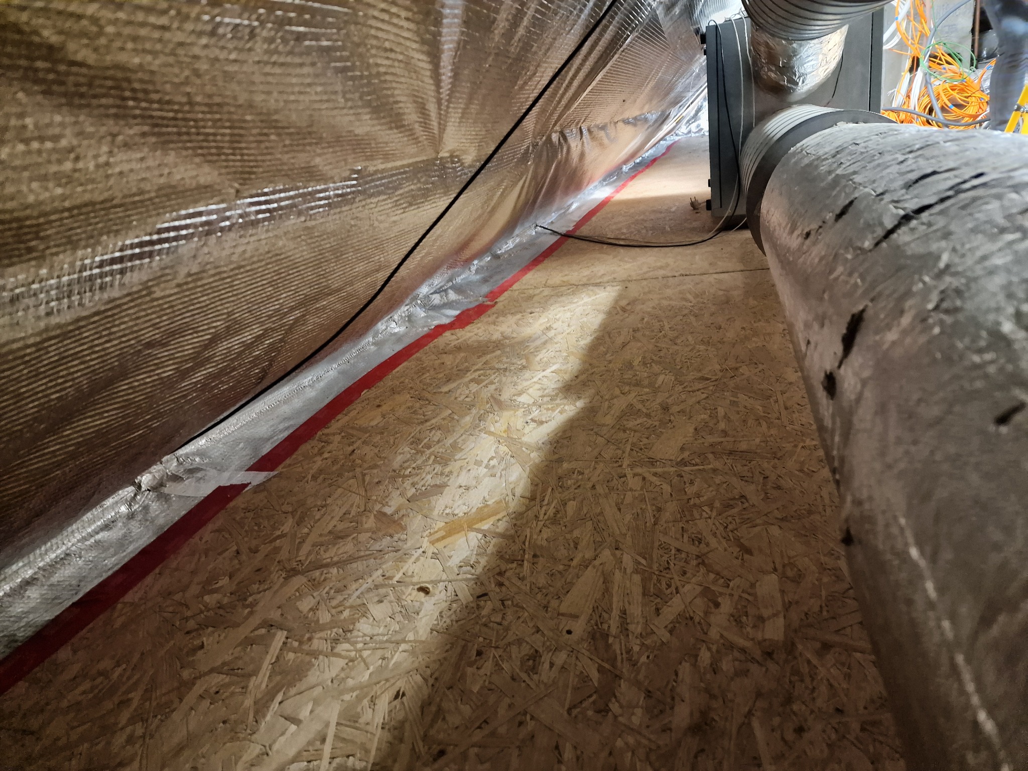 Skośny widok na poddasze z folią izolacyjną z czerwoną taśmą, drewnianą podłogą i fragmentem rury wentylacyjnej. Widoczne kable i instalacje.