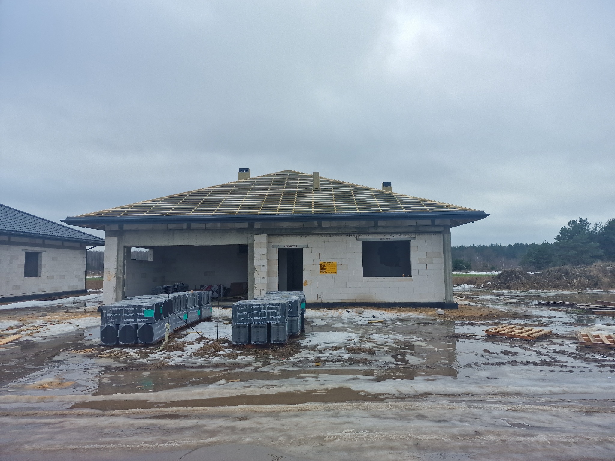 Dom w stanie surowym otwartym z widoczną konstrukcją dachu i stosem dachówek CREATON. Na pierwszym planie błoto i resztki śniegu. Pochmurne niebo.