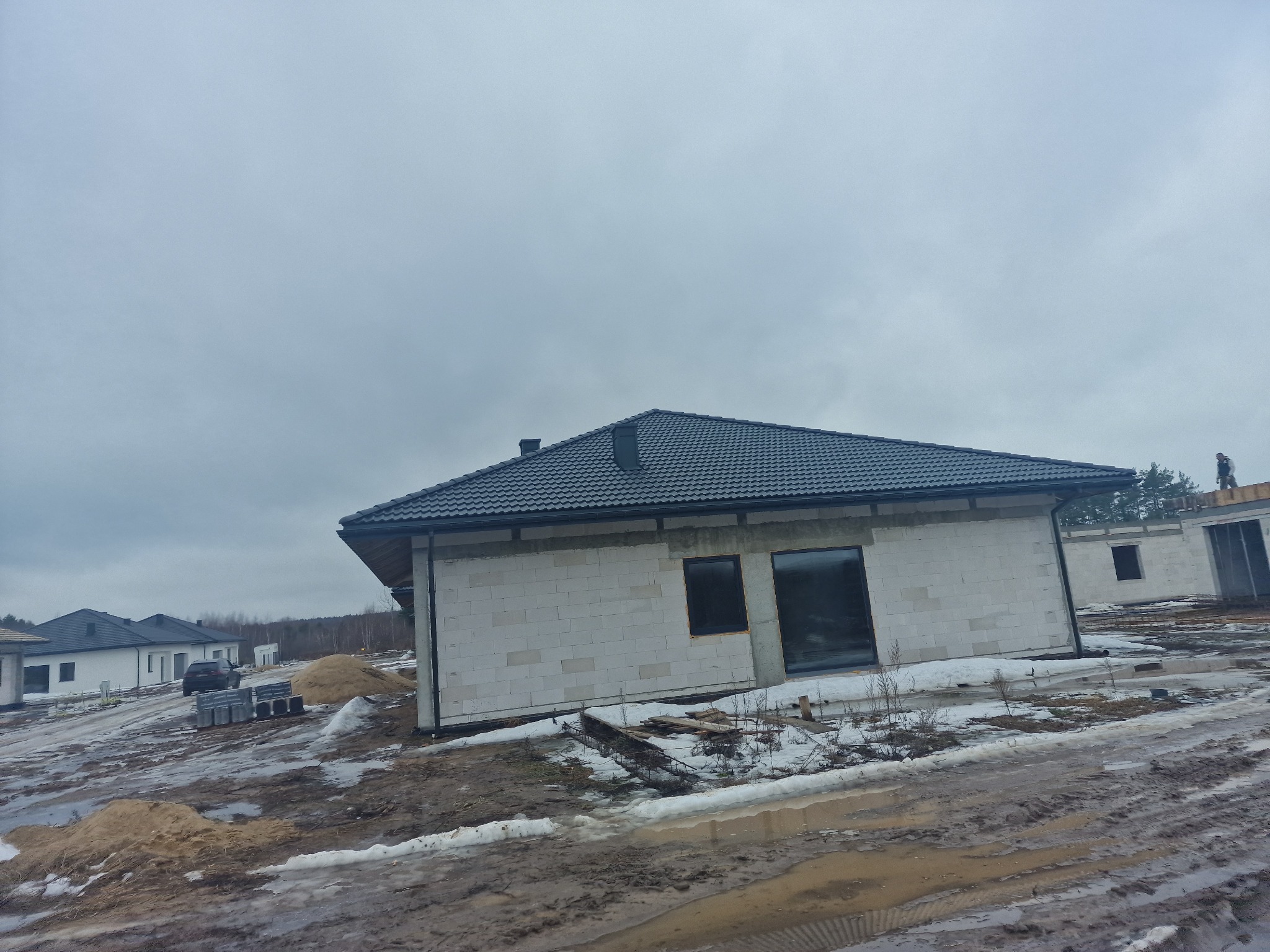 Nowoczesny dom jednorodzinny w trakcie budowy, z ciemną dachówką i białymi ścianami z bloczków, na zasypanym śniegiem terenie. Widoczny fragment dachu sąsiedniego budynku.