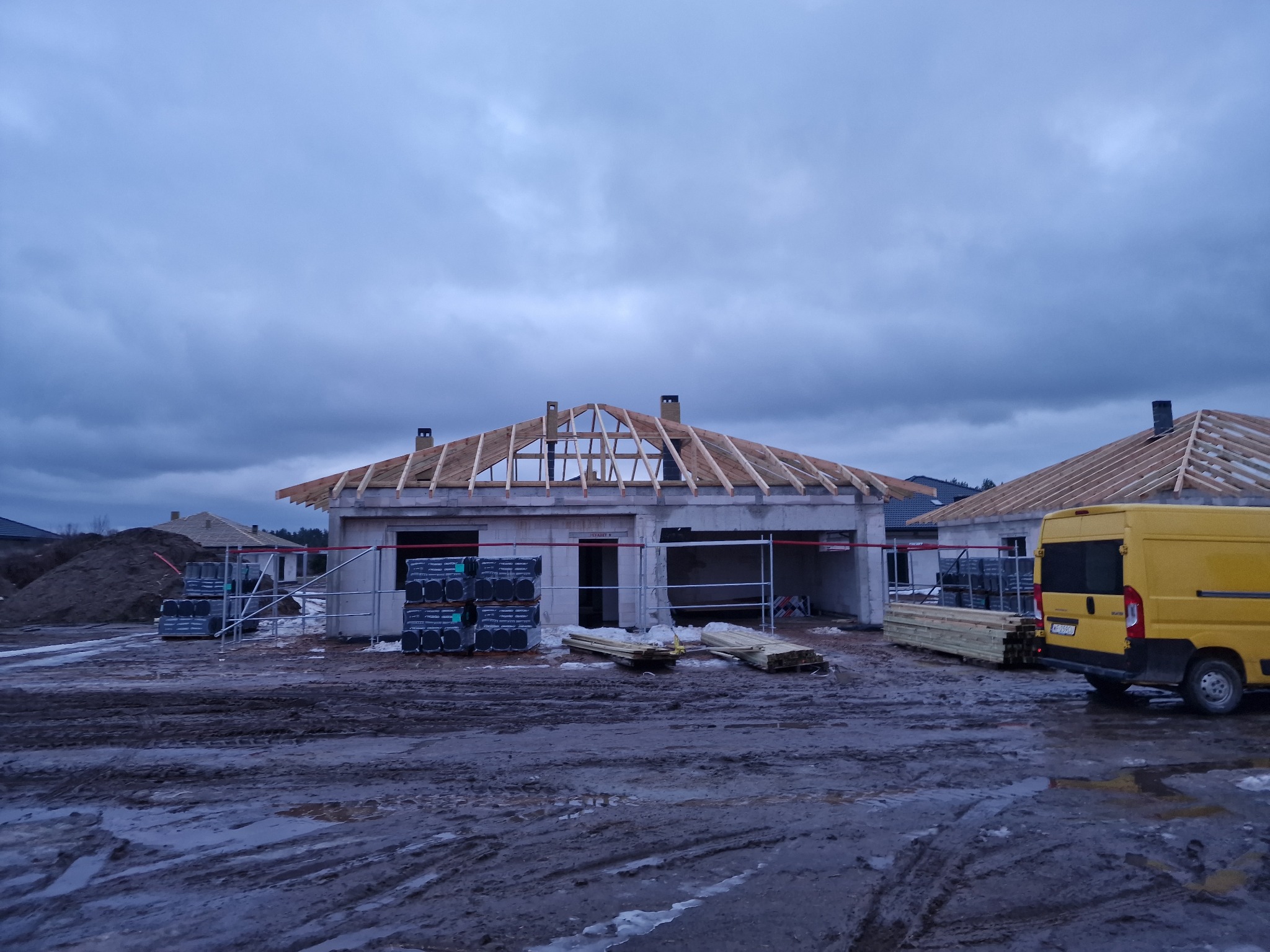 Budowa domu w trakcie krycia dachu, widoczne krokwie i kominy. Na pierwszym planie materiały budowlane i żółty samochód dostawczy na błotnistym terenie.