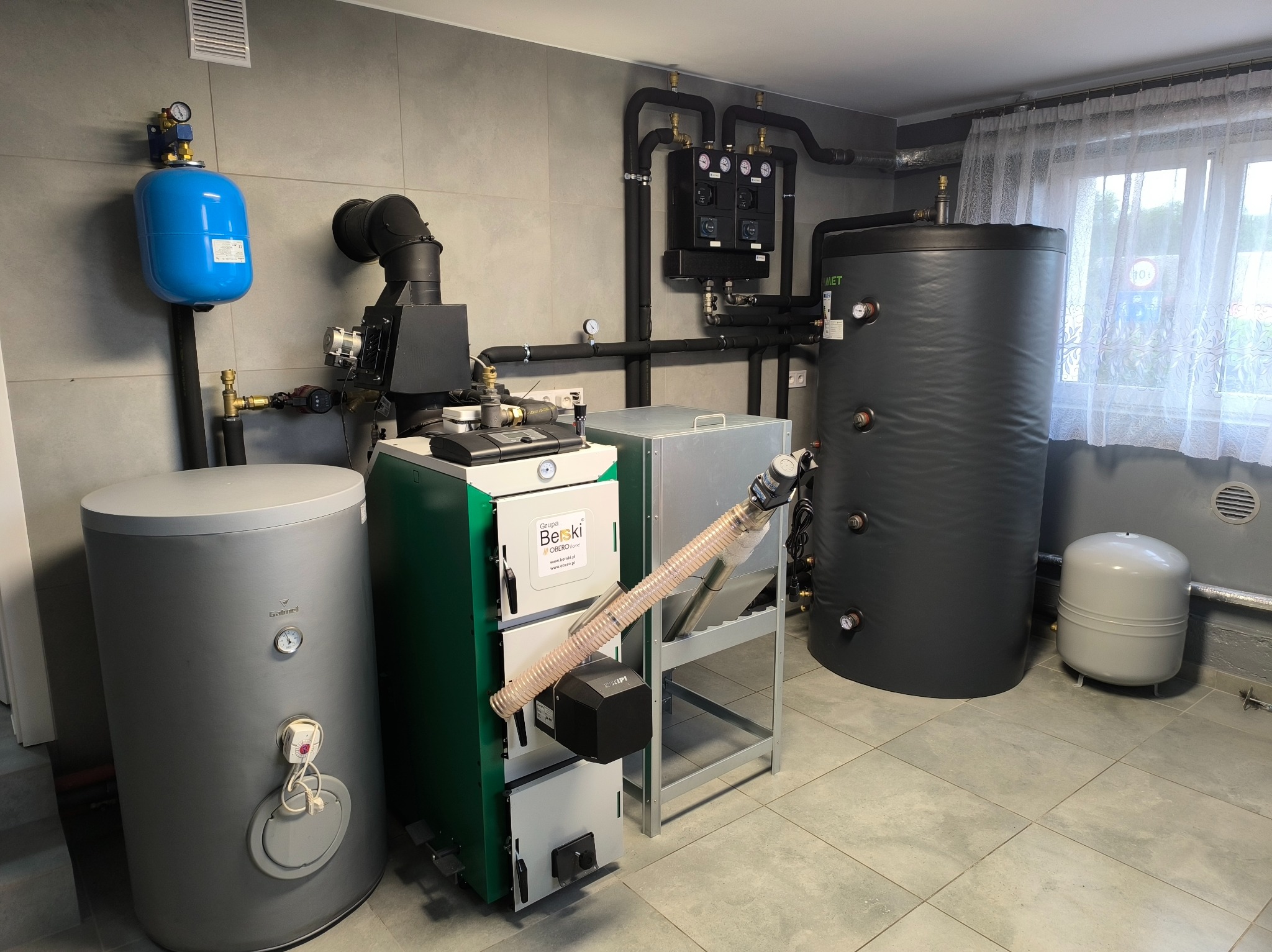 Kompletna kotłownia z piecem na ekogroszek Beski, zbiornikiem buforowym MET, zasobnikiem CWU Galmet i osprzętem. Szare płytki na ścianach i podłodze.