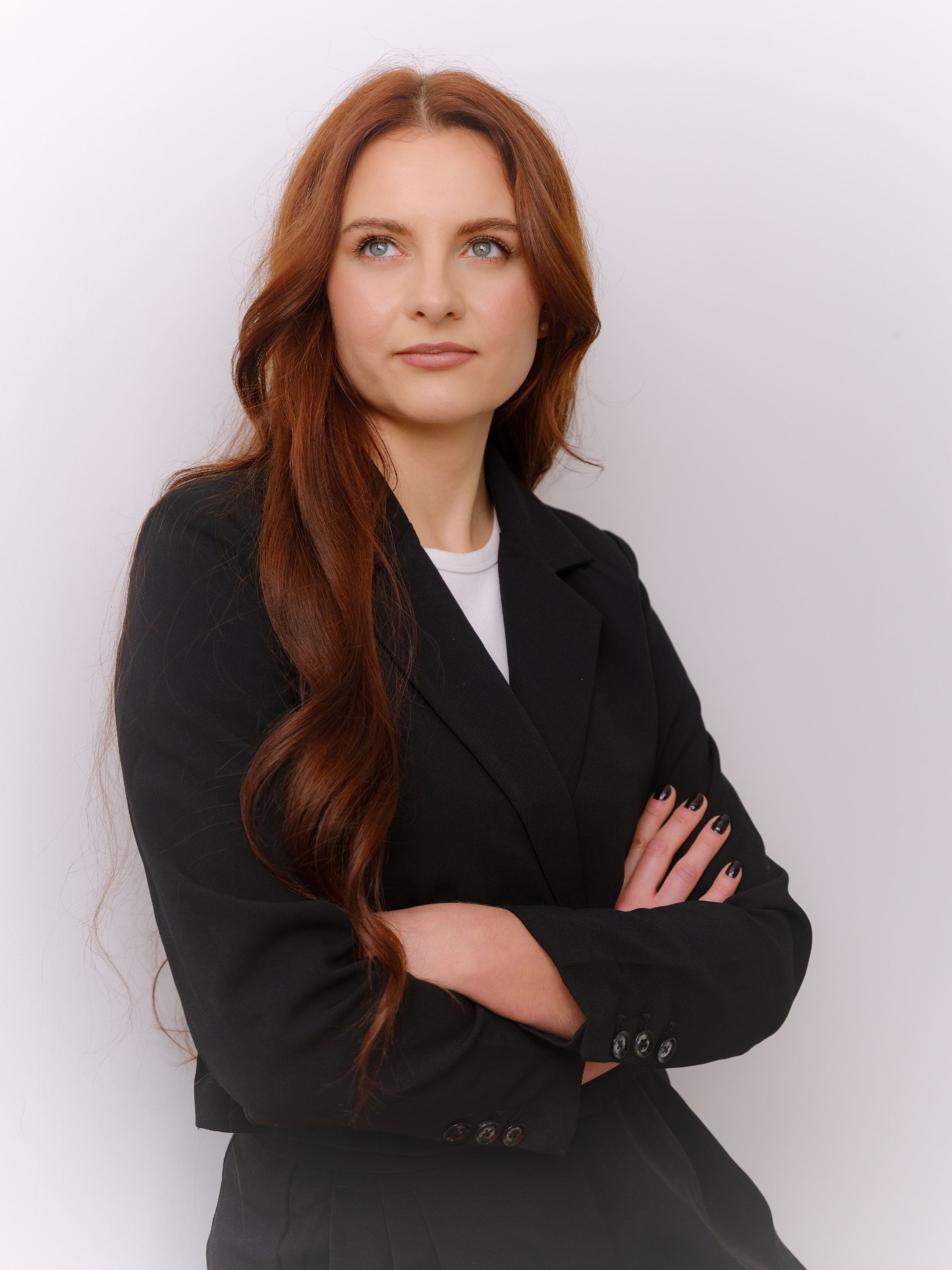 Portret kobiety w czarnym żakiecie, z rudymi włosami i skrzyżowanymi rękami, spoglądającej w górę, na jasnym tle. Profesjonalny wizerunek.