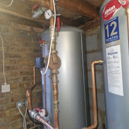 Mariusz Michalak - Instalacja z dwoma szarymi zbiornikami, miedzianymi rurami i zaworami z czerwonymi uchwytami na tle ceglanej ściany i drewnianego sufitu. Widoczny manometr i etykiety zbiorników.