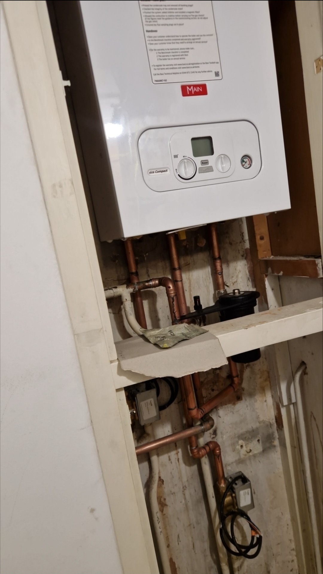 Biały piec gazowy marki Main, zamontowany na ścianie z widocznymi miedzianymi rurami instalacji. Widoczne elementy instalacji i fragment ściany.