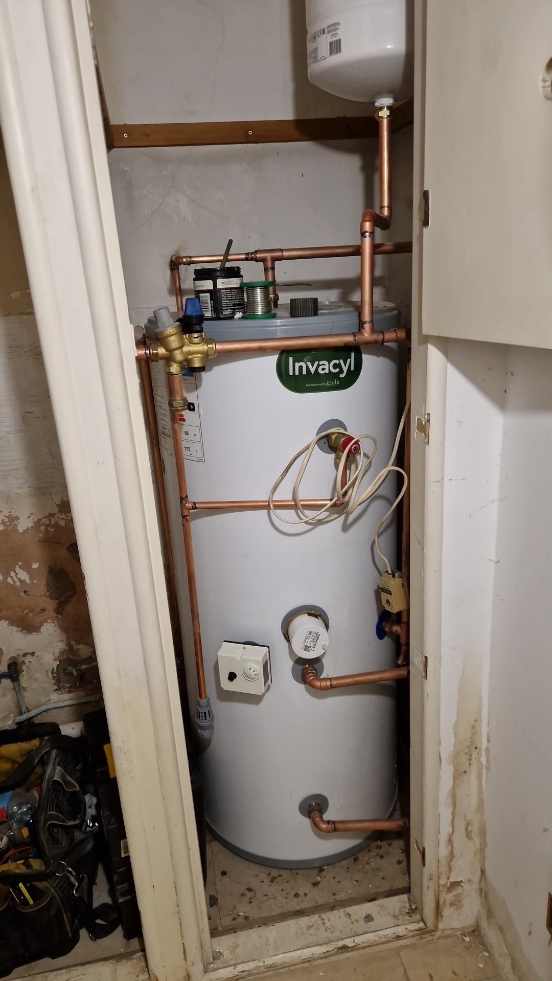 Bojler Invacyl z miedzianą instalacją w ciasnej wnęce, widoczne narzędzia hydrauliczne i ślady wilgoci na ścianach. Ujęcie z bliska, podkreślające detale instalacji.