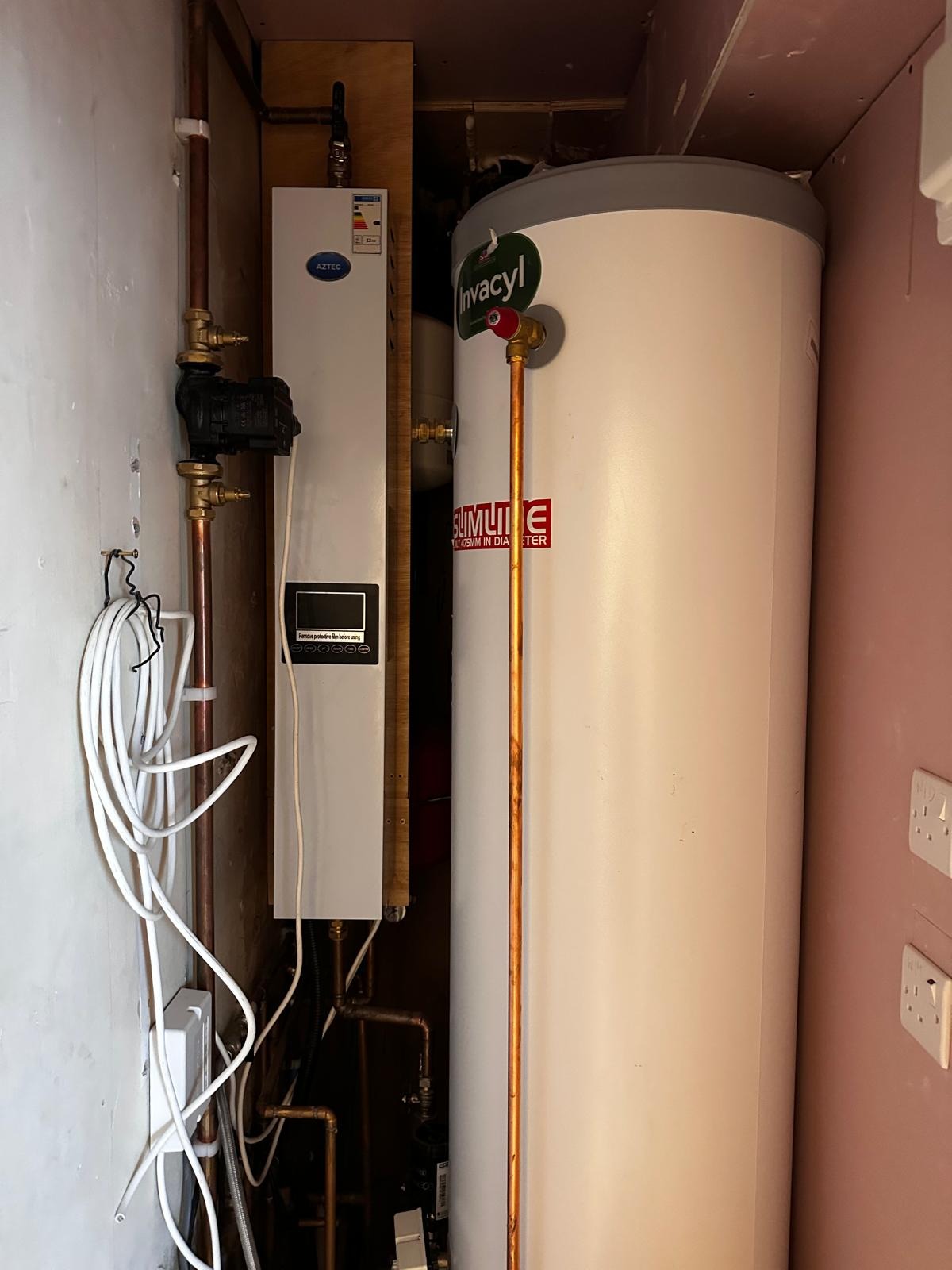 Biały, pionowy zbiornik na wodę Invacyl i piec grzewczy Aztec z miedzianymi rurami w ciasnej kotłowni. Widoczne przewody i gniazdka elektryczne.