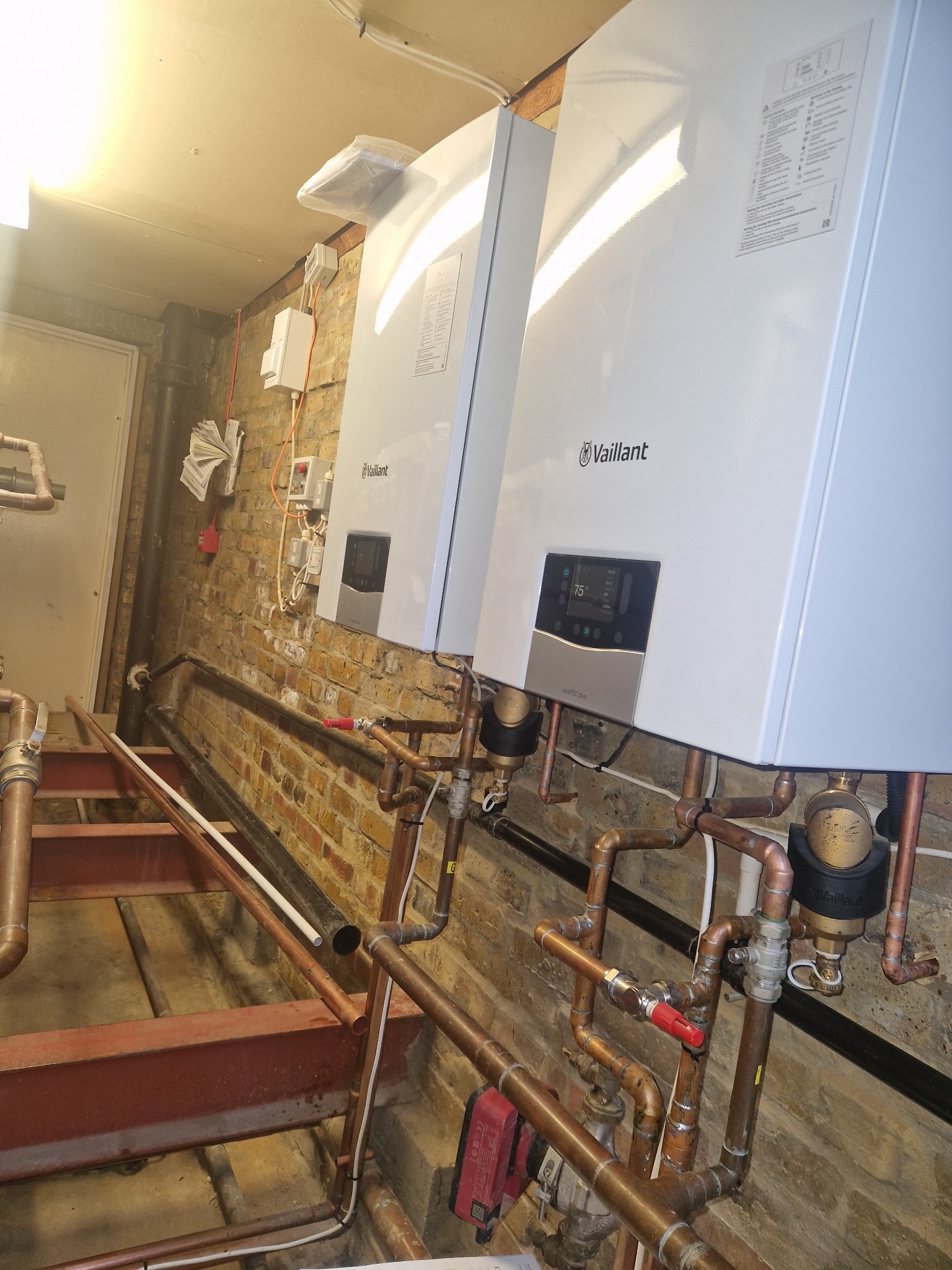 Dwa białe, wiszące piece gazowe Vaillant z podłączoną miedzianą instalacją na tle ceglanej ściany. Widoczne rury i elementy instalacji grzewczej.