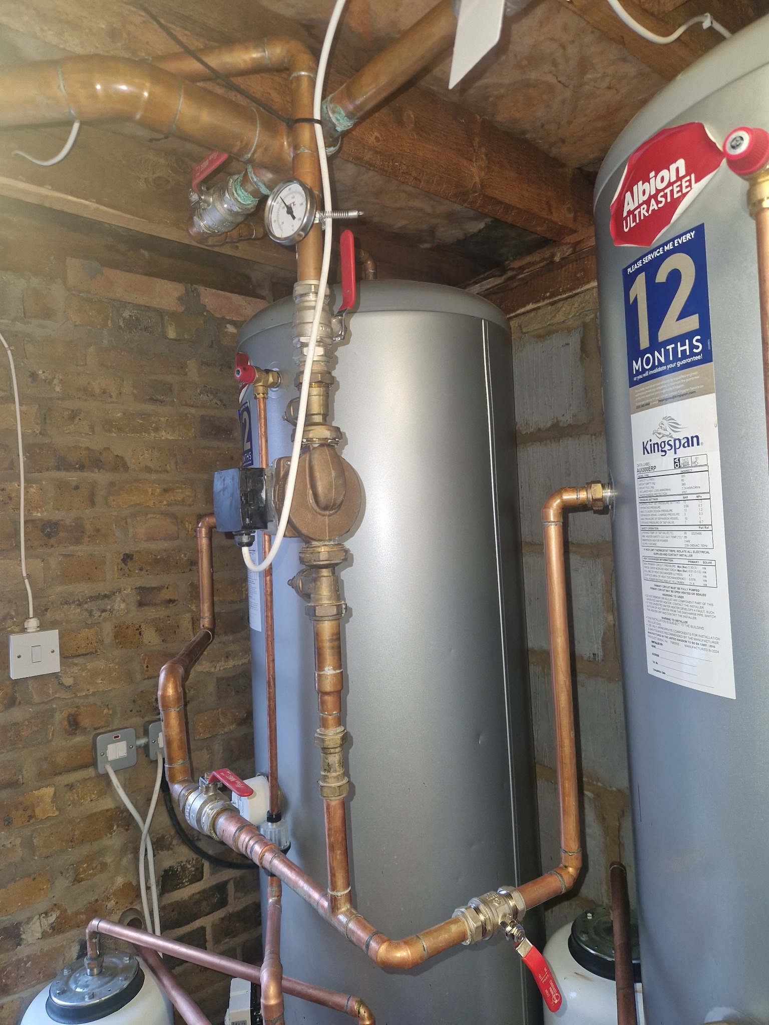 Instalacja z dwoma szarymi zbiornikami, miedzianymi rurami i zaworami z czerwonymi uchwytami na tle ceglanej ściany i drewnianego sufitu. Widoczny manometr i etykiety zbiorników.
