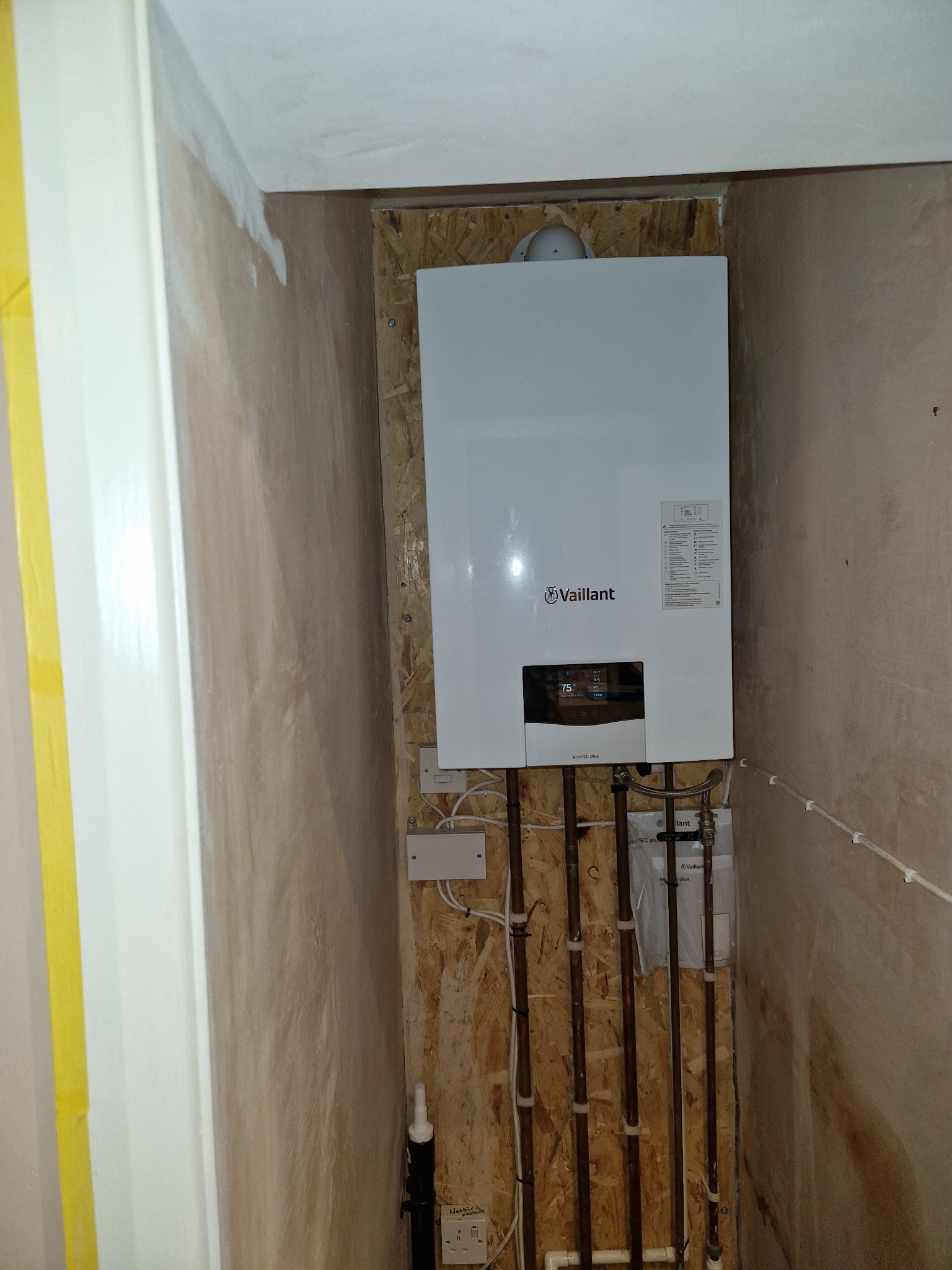 Biały, wiszący piec gazowy Vaillant zamontowany na płycie OSB w surowym wnętrzu. Widoczne miedziane rury instalacji. Ściany w trakcie wykończenia.