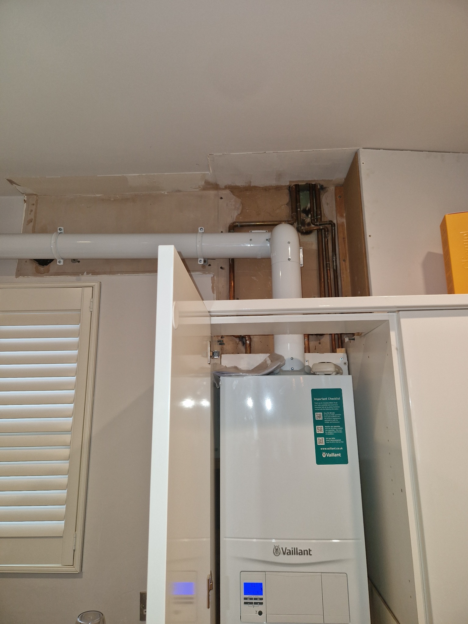 Biały piec gazowy Vaillant zamontowany w szafce kuchennej, widoczne rury instalacji i fragment okna z żaluzjami. Ściana w trakcie wykończenia.
