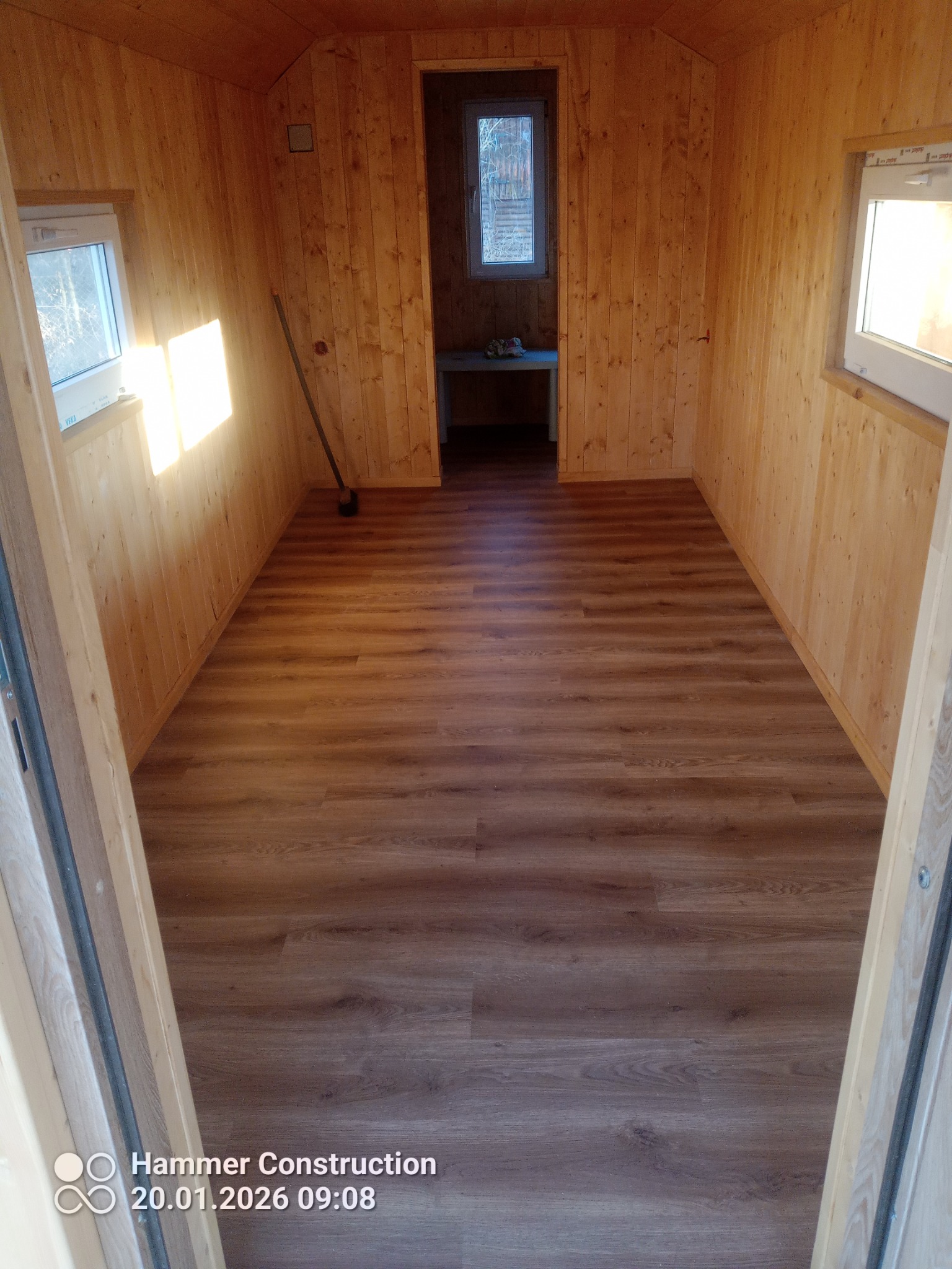 Wnętrze wykończonego pomieszczenia z drewnianą boazerią i panelami podłogowymi w odcieniach brązu. Widoczne okna i drzwi. W rogu stoi miotła.