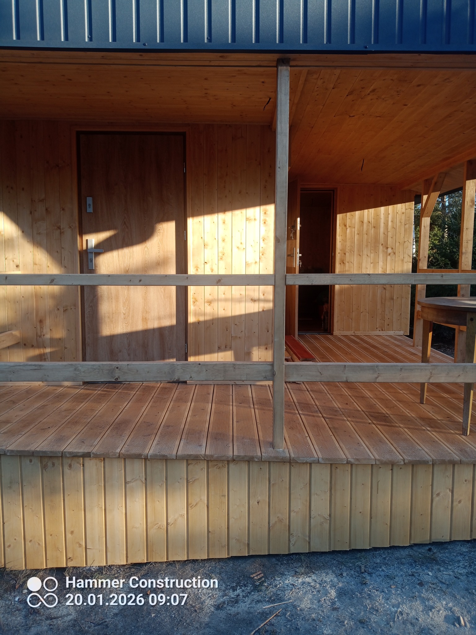 Drewniany taras z balustradą i dwoma wejściami, oświetlony ciepłym światłem słonecznym. Widoczne deski tarasowe i boazeria, metalowe zadaszenie.