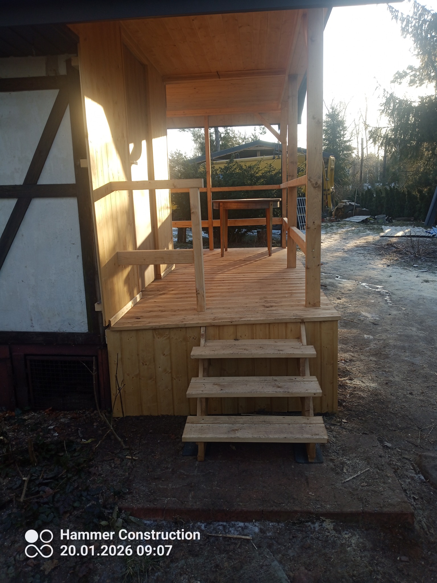 Drewniana weranda z balustradą i schodami, przylegająca do budynku z elementami muru pruskiego. Widoczne drzewa w tle, poranne słońce oświetla konstrukcję.