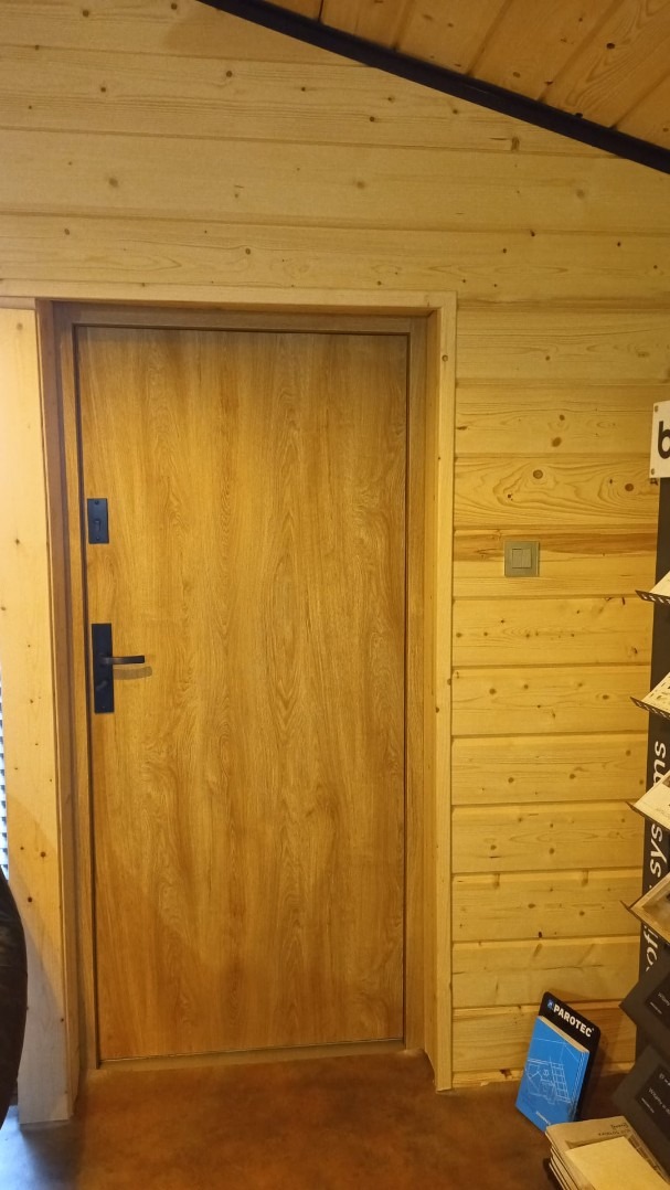 Zamknięte, drewniane drzwi wewnętrzne z czarną klamką, wbudowane w ścianę z drewnianych paneli. Widoczny fragment sufitu i podłogi. U dołu materiały informacyjne.