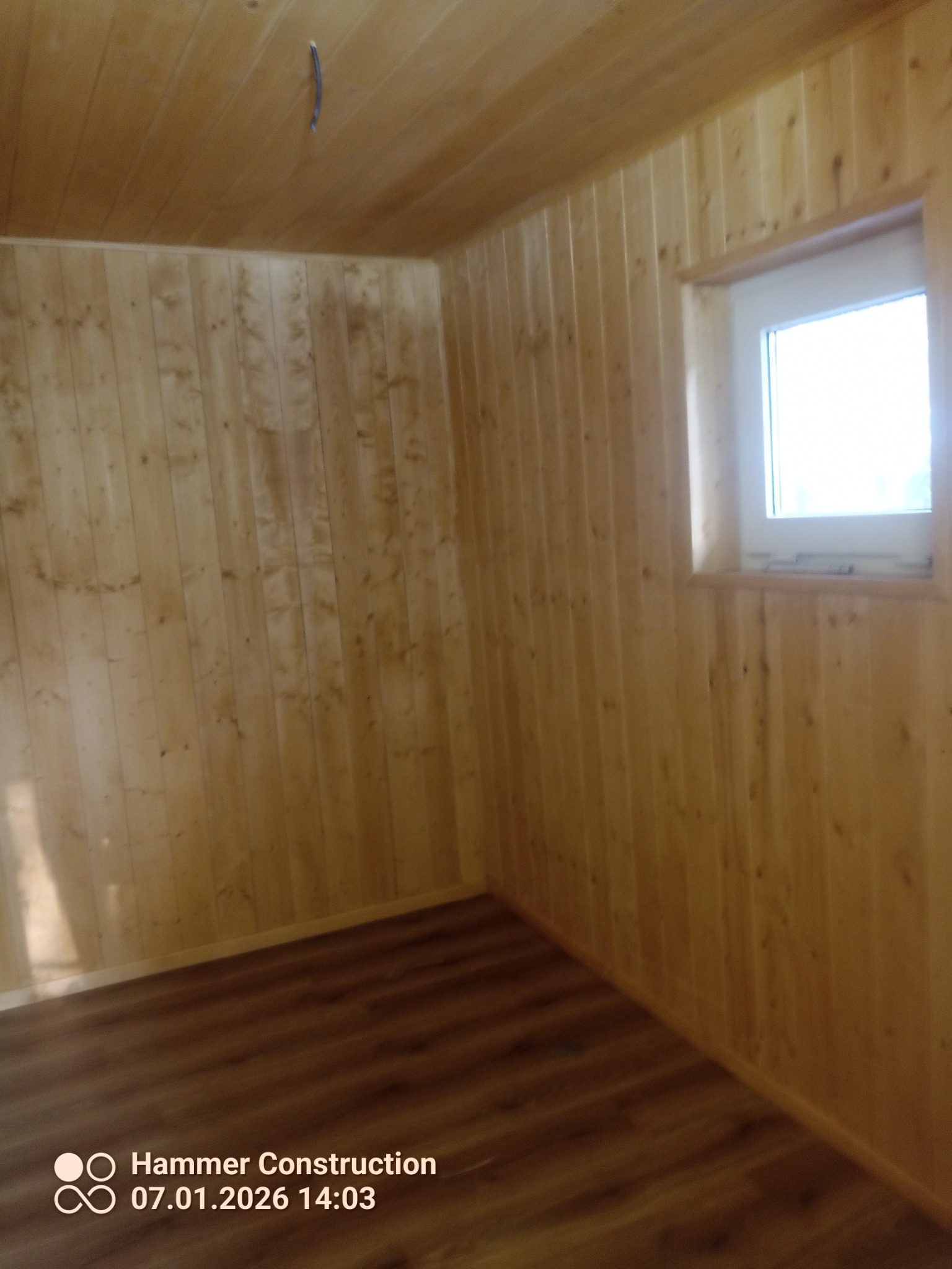 Wnętrze wykończone boazerią w odcieniach drewna, z widocznym oknem i fragmentem podłogi. Widoczny kabel wystający z sufitu.