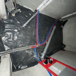 ELIASZ MARCINIAK NOWOCZESNE TECHNOLOGIE GRZEWCZE - Wnętrze w trakcie prac hydraulicznych: rury (czerwone, niebieskie, szare) prowadzone pod sufitem i wzdłuż ściany, czarna folia ochronna, stelaż podtynkowy WC.