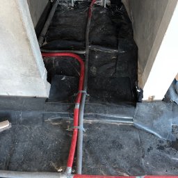 ELIASZ MARCINIAK NOWOCZESNE TECHNOLOGIE GRZEWCZE - Czerwone i szare rury instalacyjne, zabezpieczone obejmami, ułożone na czarnej folii w surowym wnętrzu. Widoczne elementy konstrukcyjne budynku.