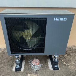 ELIASZ MARCINIAK NOWOCZESNE TECHNOLOGIE GRZEWCZE - Szara pompa ciepła Heiko zamontowana na zewnątrz budynku, widoczna kratka wentylacyjna i logo producenta. Montaż na podłożu z ziemi i kamieni.