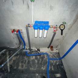 ELIASZ MARCINIAK NOWOCZESNE TECHNOLOGIE GRZEWCZE - Instalacja filtrów wody z niebieskimi rurami w surowym wnętrzu. Widoczne zawory, rury szare i niebieskie, oraz surowy beton na ścianach i podłodze.