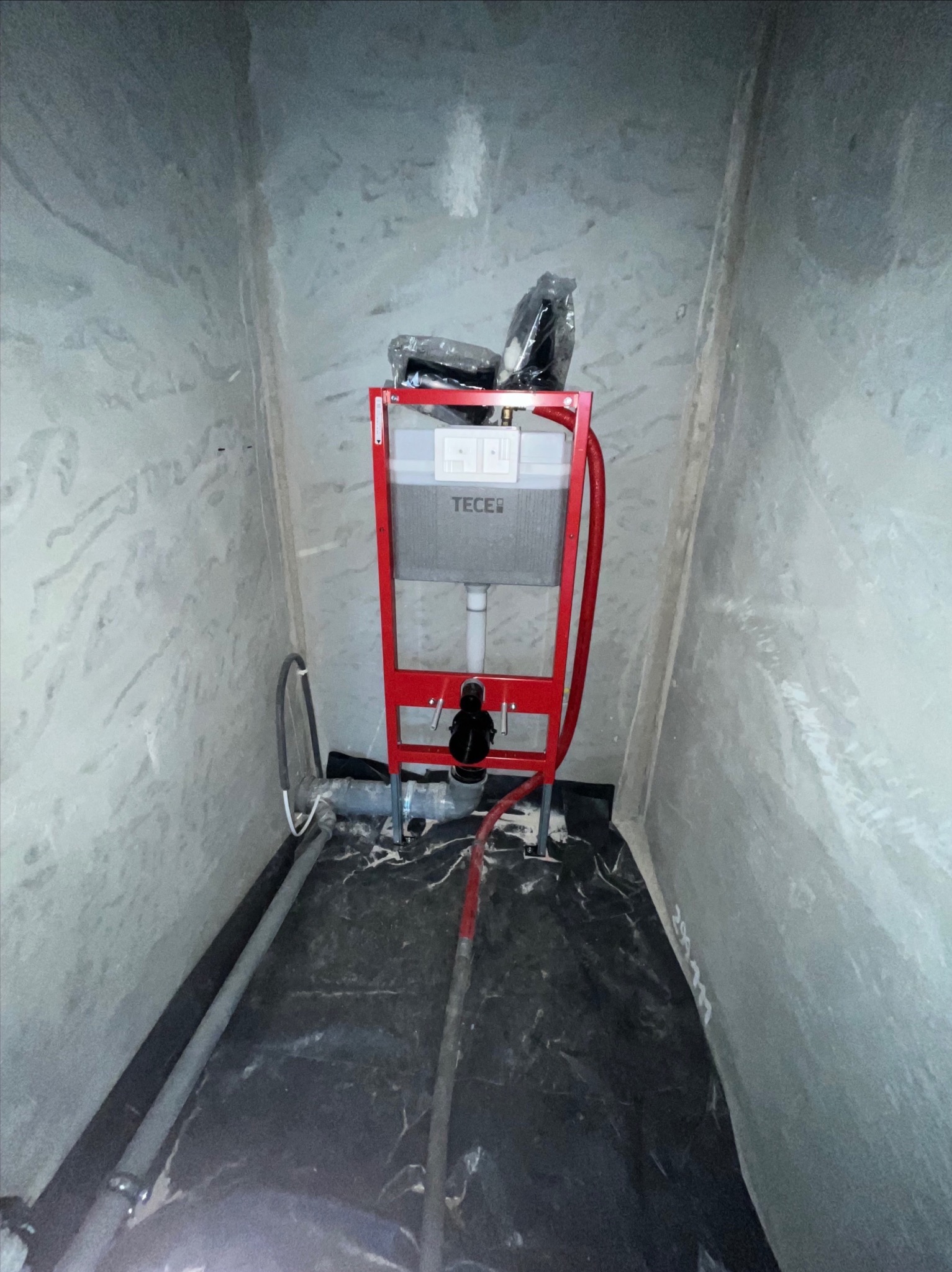 Stelaż podtynkowy WC marki Tece w trakcie montażu, widoczne rury doprowadzające wodę i odpływ. Ściany surowe, w trakcie wykończenia. Podłoga zabezpieczona folią.