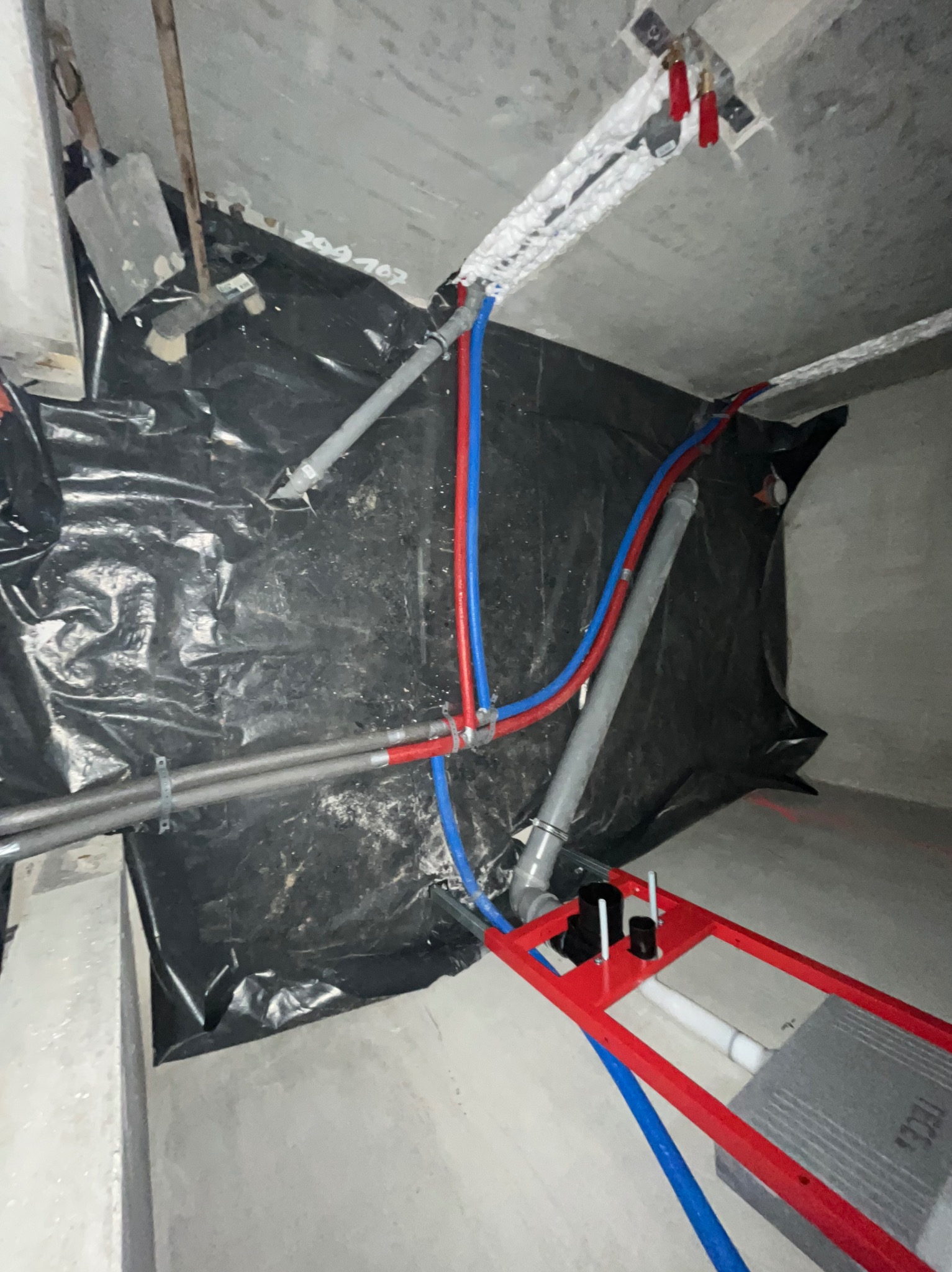 Wnętrze w trakcie prac hydraulicznych: rury (czerwone, niebieskie, szare) prowadzone pod sufitem i wzdłuż ściany, czarna folia ochronna, stelaż podtynkowy WC.