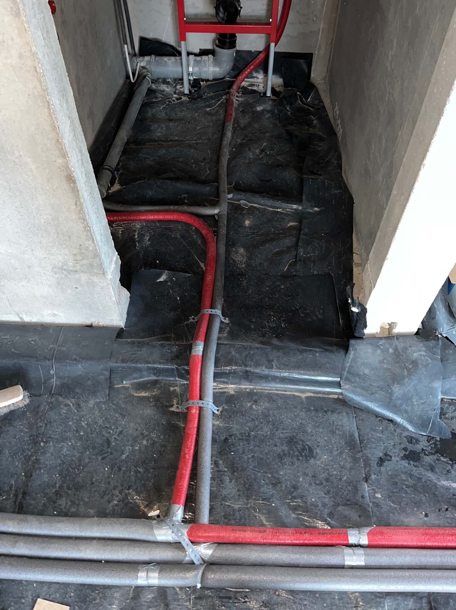 Czerwone i szare rury instalacyjne, zabezpieczone obejmami, ułożone na czarnej folii w surowym wnętrzu. Widoczne elementy konstrukcyjne budynku.