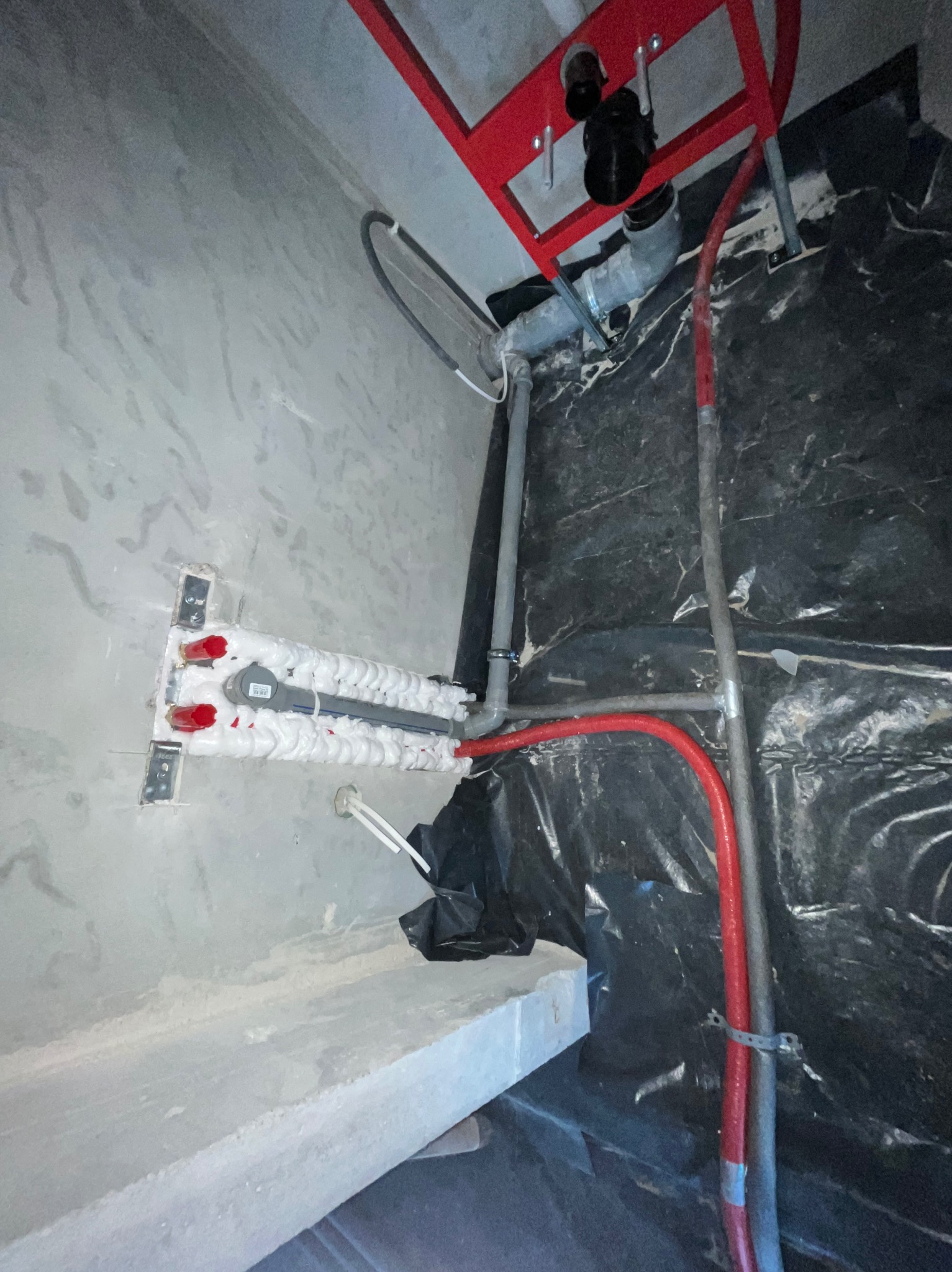 Szara ściana z instalacją wodną w izolacji piankowej, czerwone rury i stelaż podtynkowy WC na czarnej folii. Widoczne elementy montażowe i fragment betonowej konstrukcji.