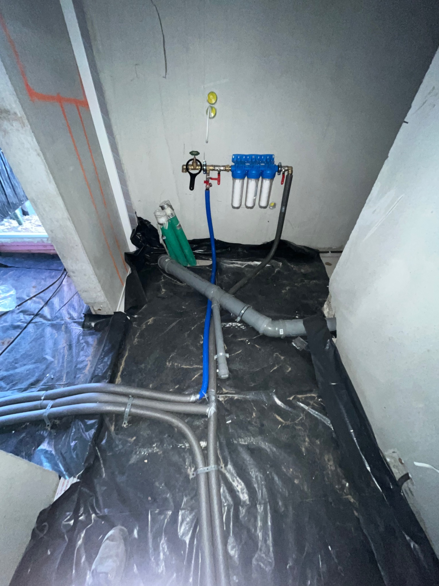Instalacja filtrów wody z rurami w pomieszczeniu z folią ochronną na podłodze. Widoczne elementy instalacji hydraulicznej i filtrów.