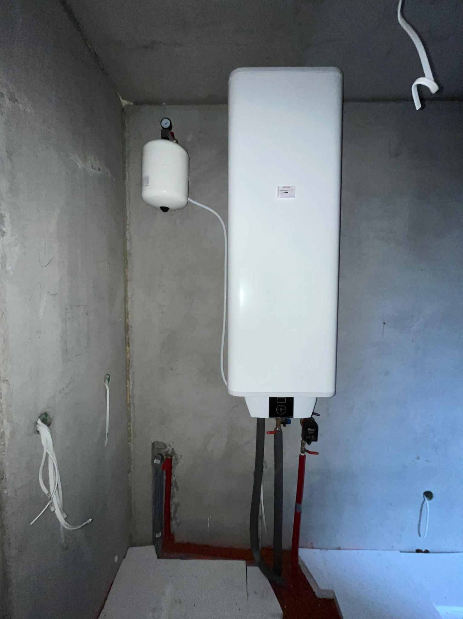 Biały, wiszący bojler elektryczny z podłączonymi rurami w surowym pomieszczeniu z betonowymi ścianami i przygotowaną instalacją. Widoczny zbiornik wyrównawczy.