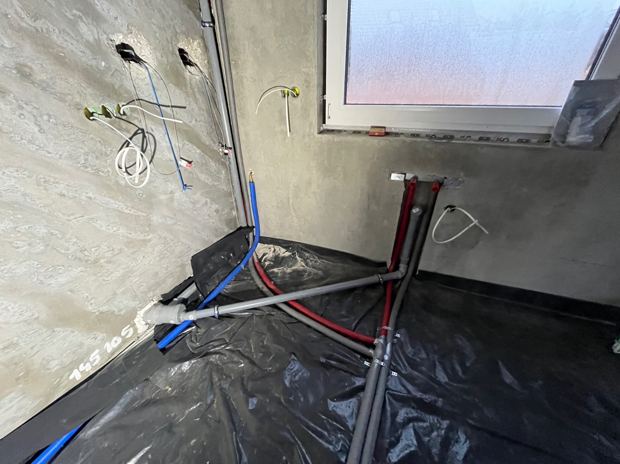 Fragment instalacji wodnej i grzewczej w surowym wnętrzu. Rury z tworzyw sztucznych w kolorach szarym, czerwonym i niebieskim ułożone na czarnej folii. Widoczne przewody elektryczne.