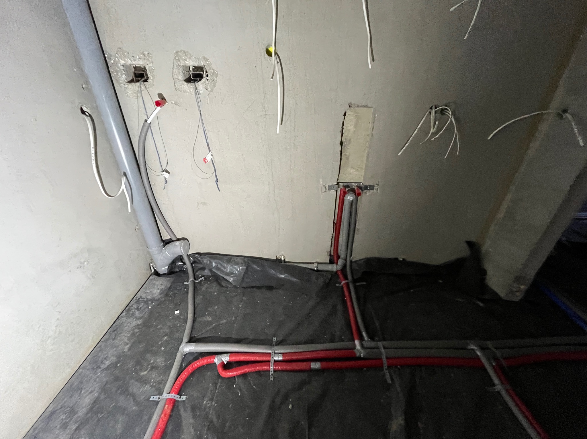Surowy stan instalacji: szare rury kanalizacyjne i czerwone rury grzewcze zamocowane do podłoża, wystające kable elektryczne ze ściany w trakcie prac wykończeniowych.