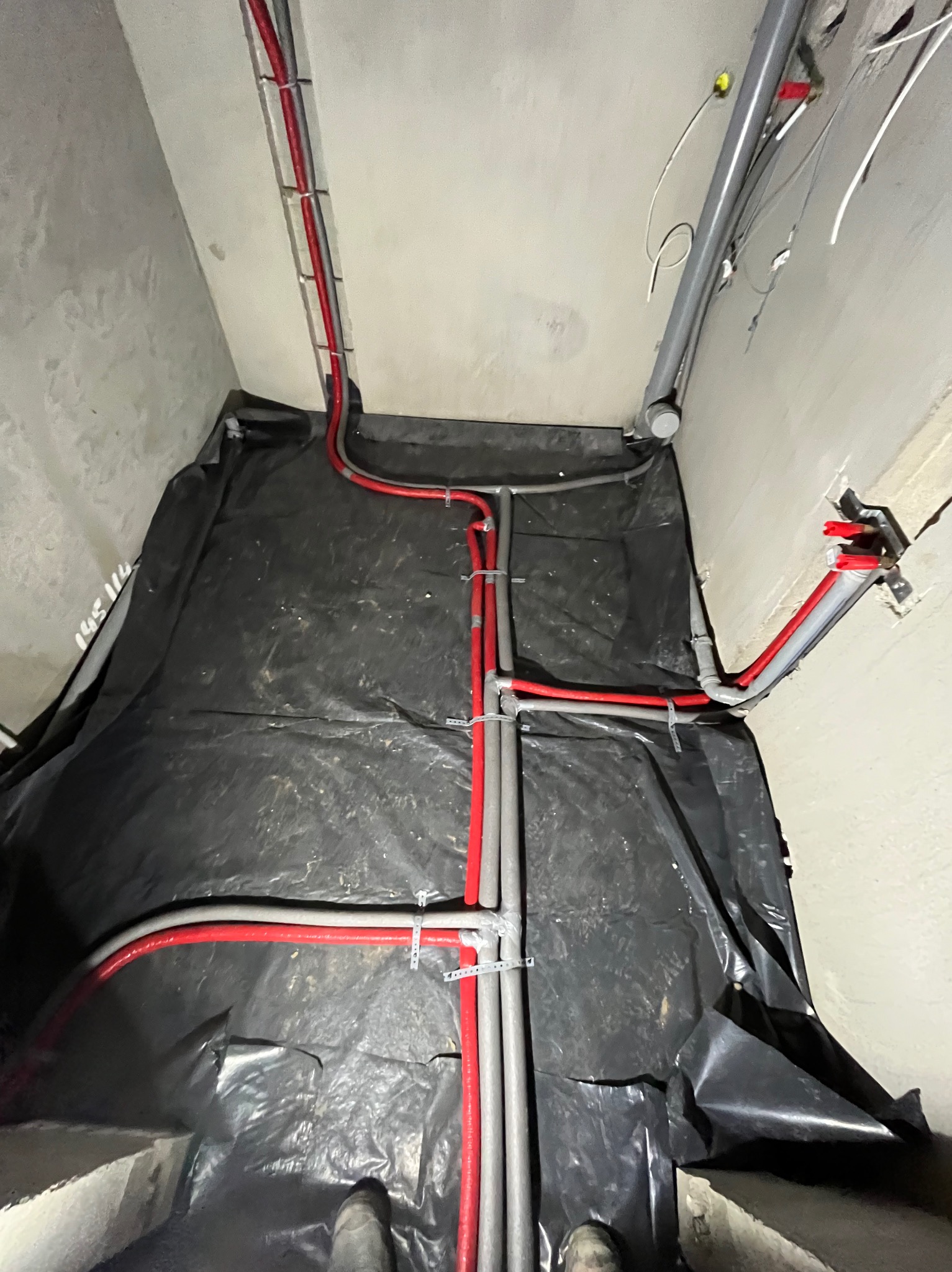 Szare i czerwone rury instalacyjne prowadzone wzdłuż ścian i pod sufitem, zabezpieczone folią. Widoczne fragmenty butów osoby wykonującej prace.