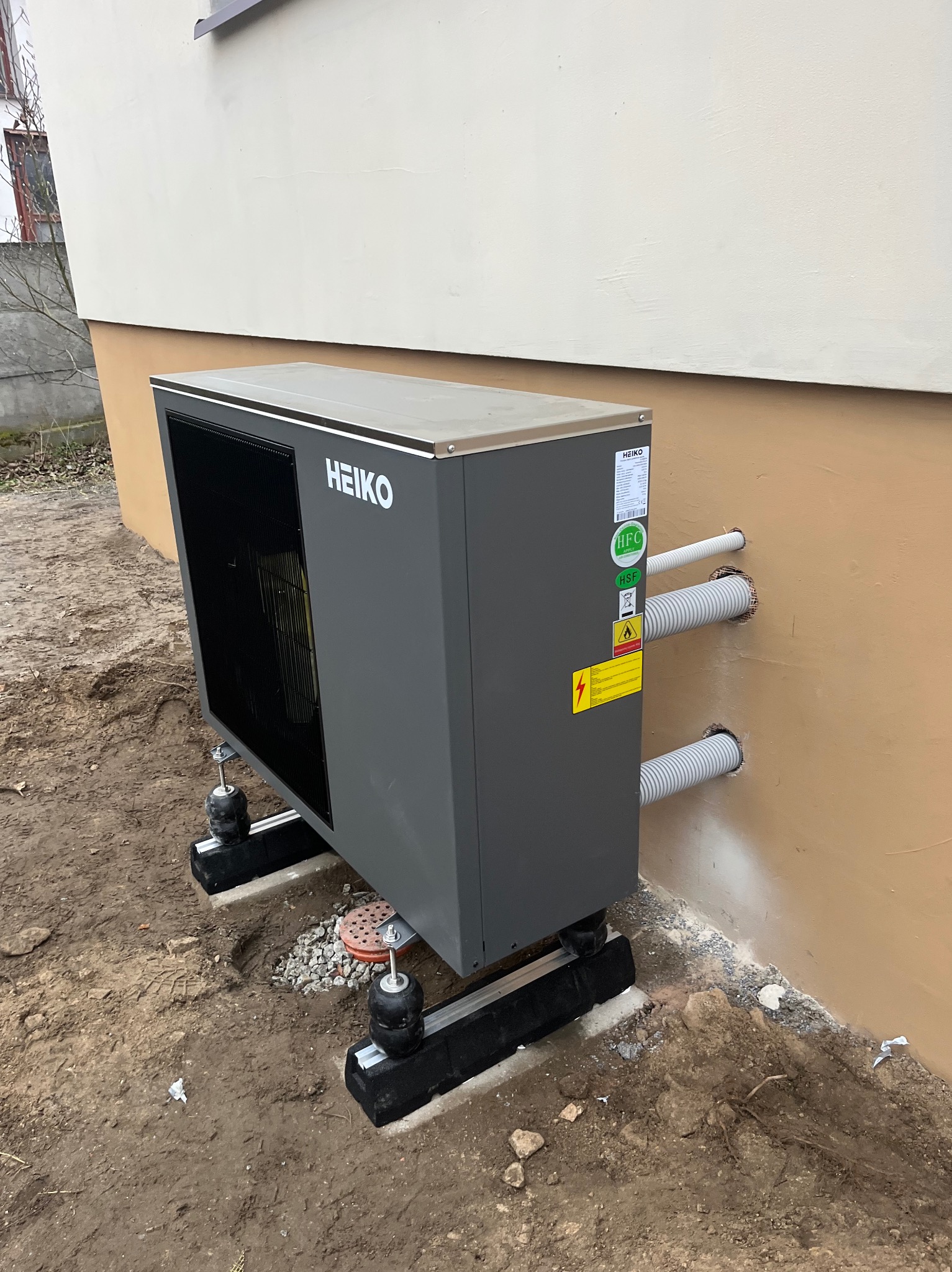 Szara pompa ciepła marki HEIKO zamontowana na zewnątrz budynku z widocznymi rurami wchodzącymi w ścianę. Widoczne elementy montażowe i teren wokół.