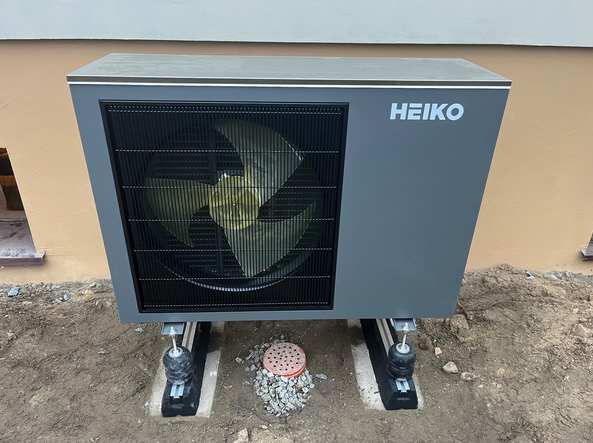 Szara pompa ciepła Heiko zamontowana na zewnątrz budynku, widoczna kratka wentylacyjna i logo producenta. Montaż na podłożu z ziemi i kamieni.
