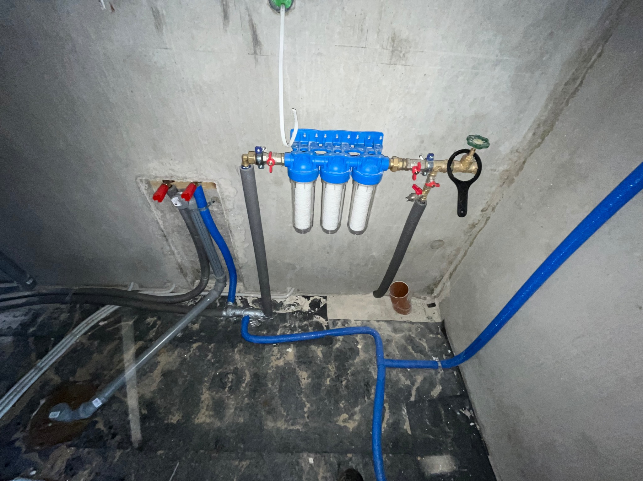 Instalacja filtrów wody z niebieskimi rurami w surowym wnętrzu. Widoczne zawory, rury szare i niebieskie, oraz surowy beton na ścianach i podłodze.