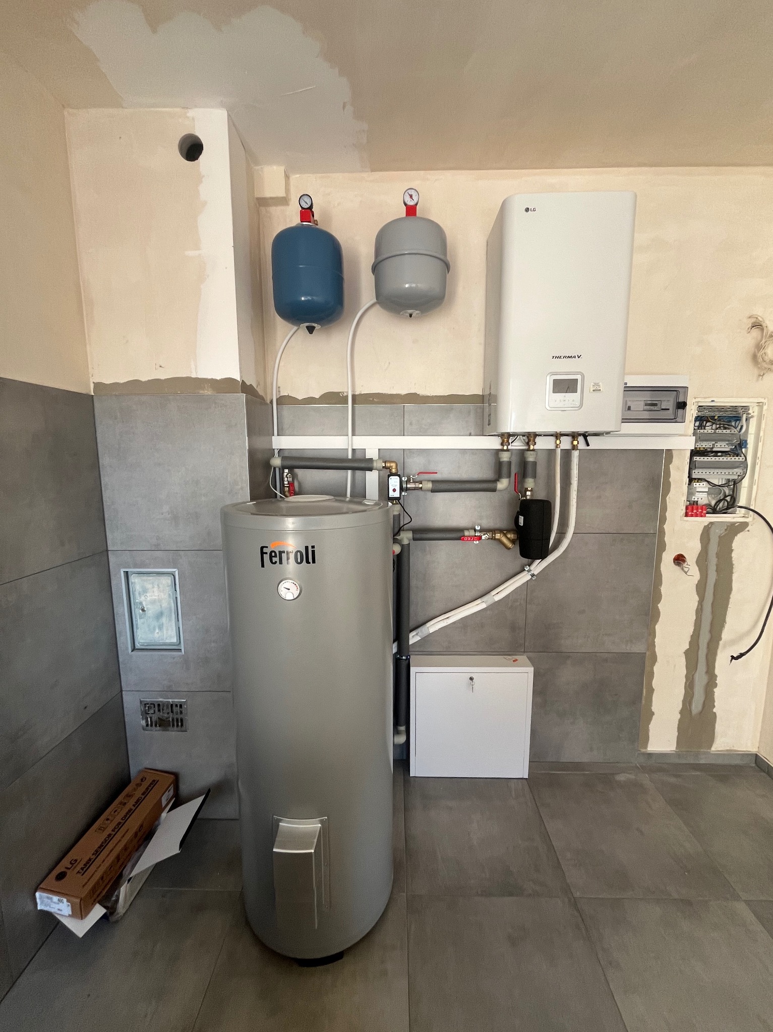 Szary bojler Ferroli, biały piec gazowy LG i dwa zbiorniki wyrównawcze na tle szarych płytek i fragmentów surowej ściany. Widoczne rury i instalacja elektryczna.