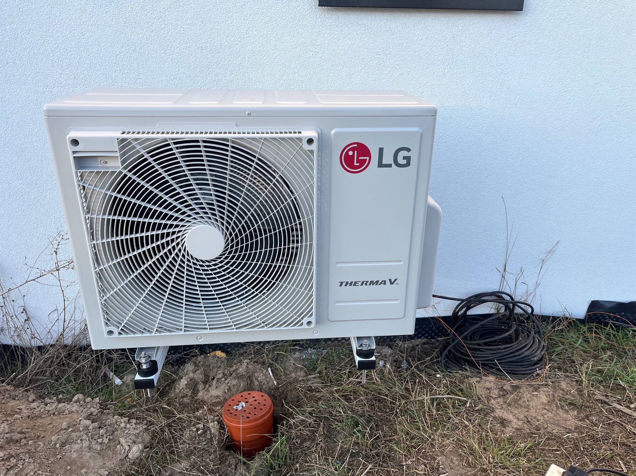 Biała pompa ciepła LG Therma V zamontowana przy ścianie budynku, widoczne przewody i elementy instalacji na trawniku. Ujęcie z bliska, skupione na detalu montażu.