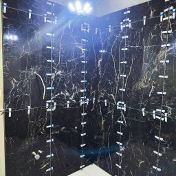 GWARANT PROBUD - Montaż czarnych płytek imitujących marmur w łazience z użyciem systemu poziomowania. Widoczne białe klipsy i kliny. Oświetlenie sufitowe.